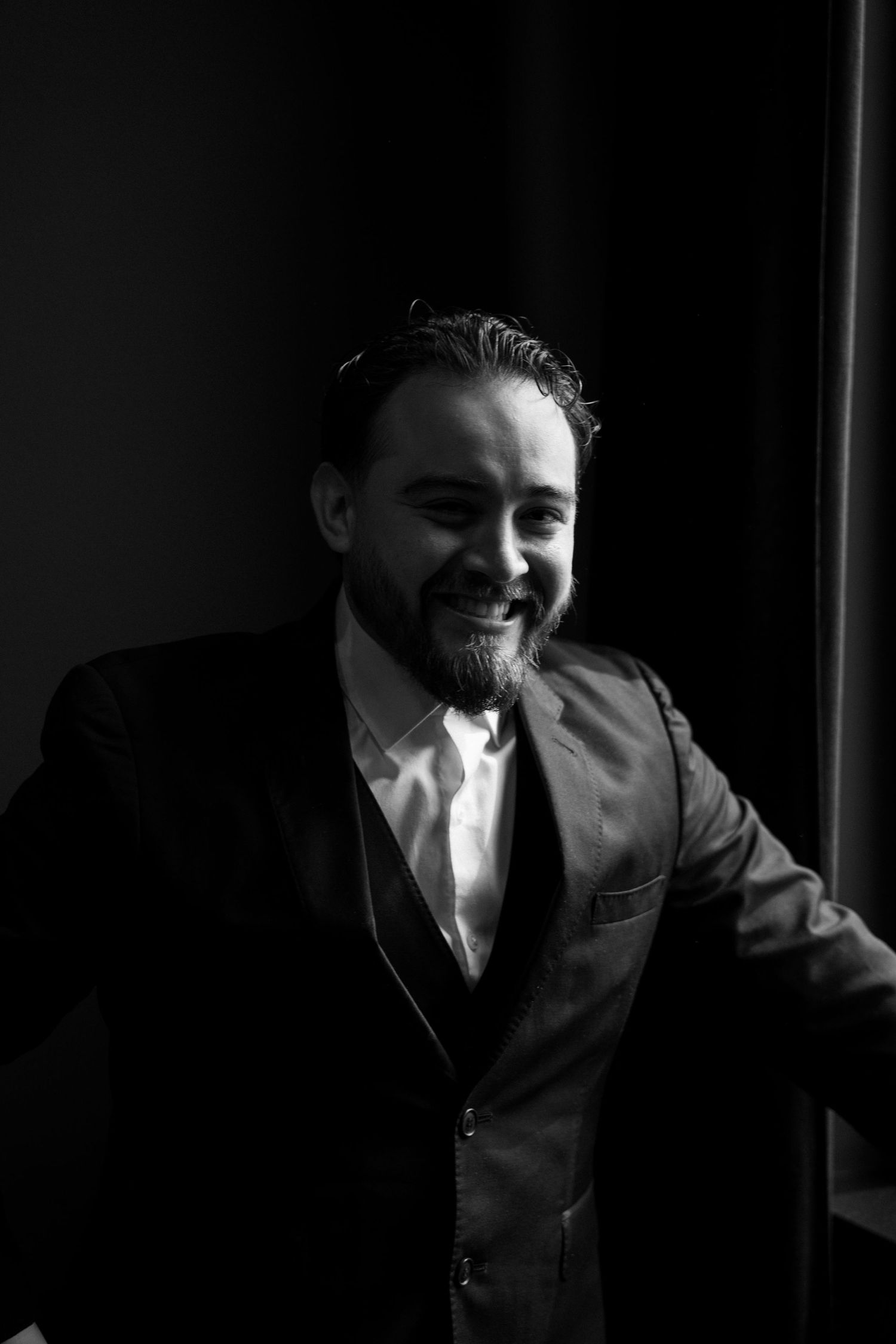 Black and white portrait of smiling bearded groom in dark suit leaning against wall.