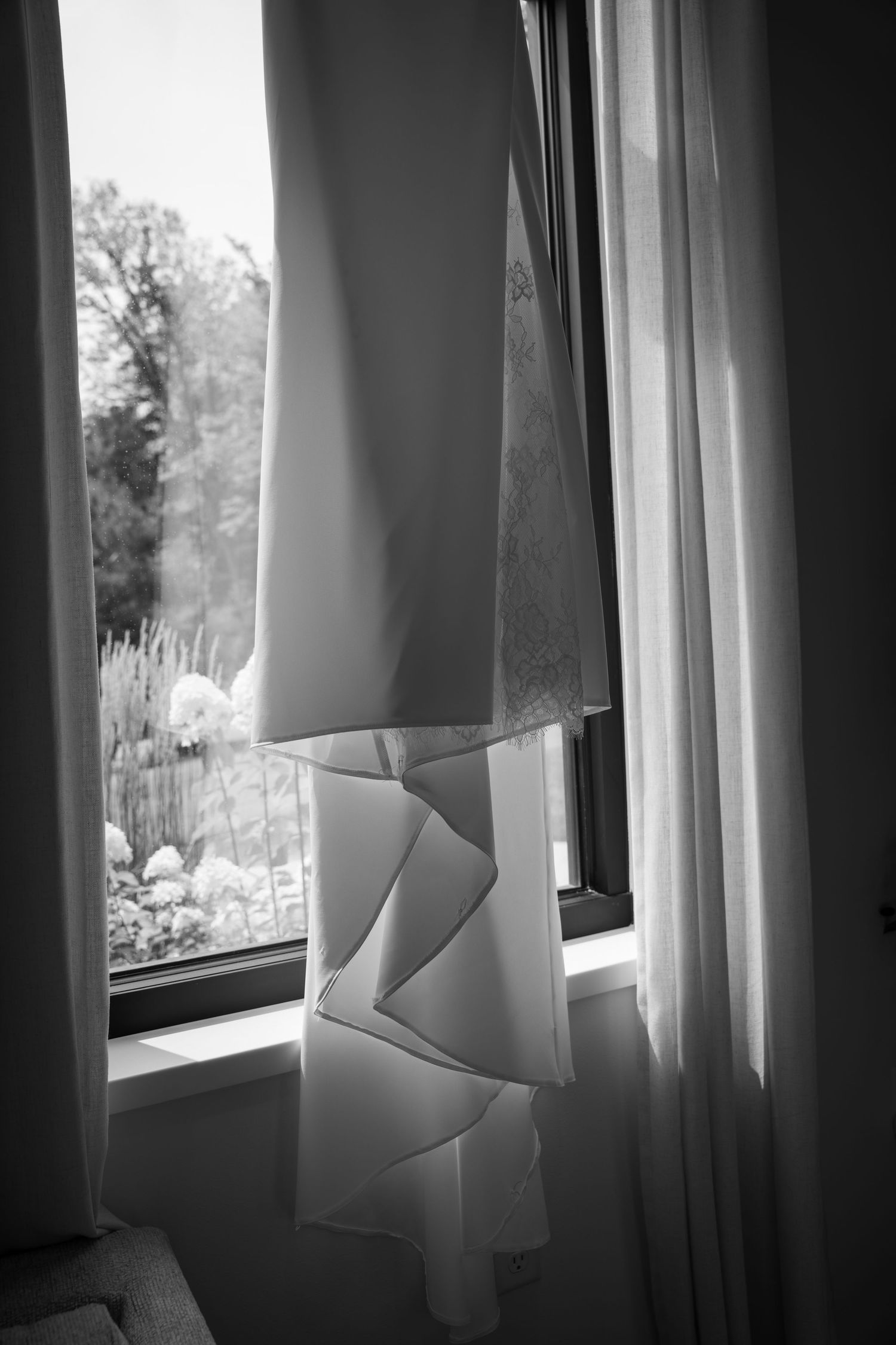 Black and white photo of wedding dress hanging in window with garden view and soft natural light.