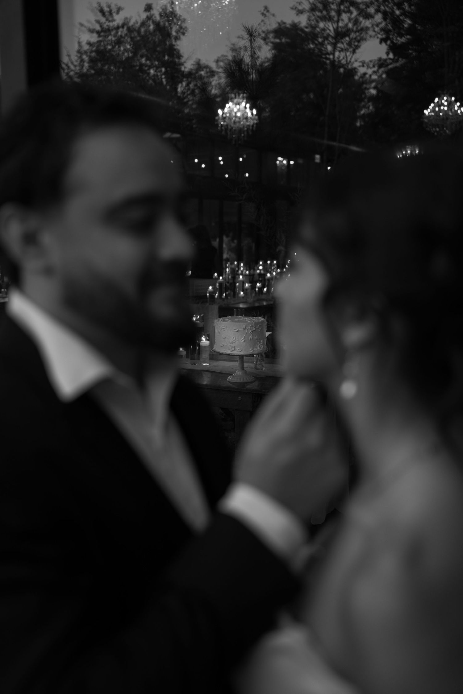 Artistic black and white photo of bride and groom with a wedding cake softly blurred in background.