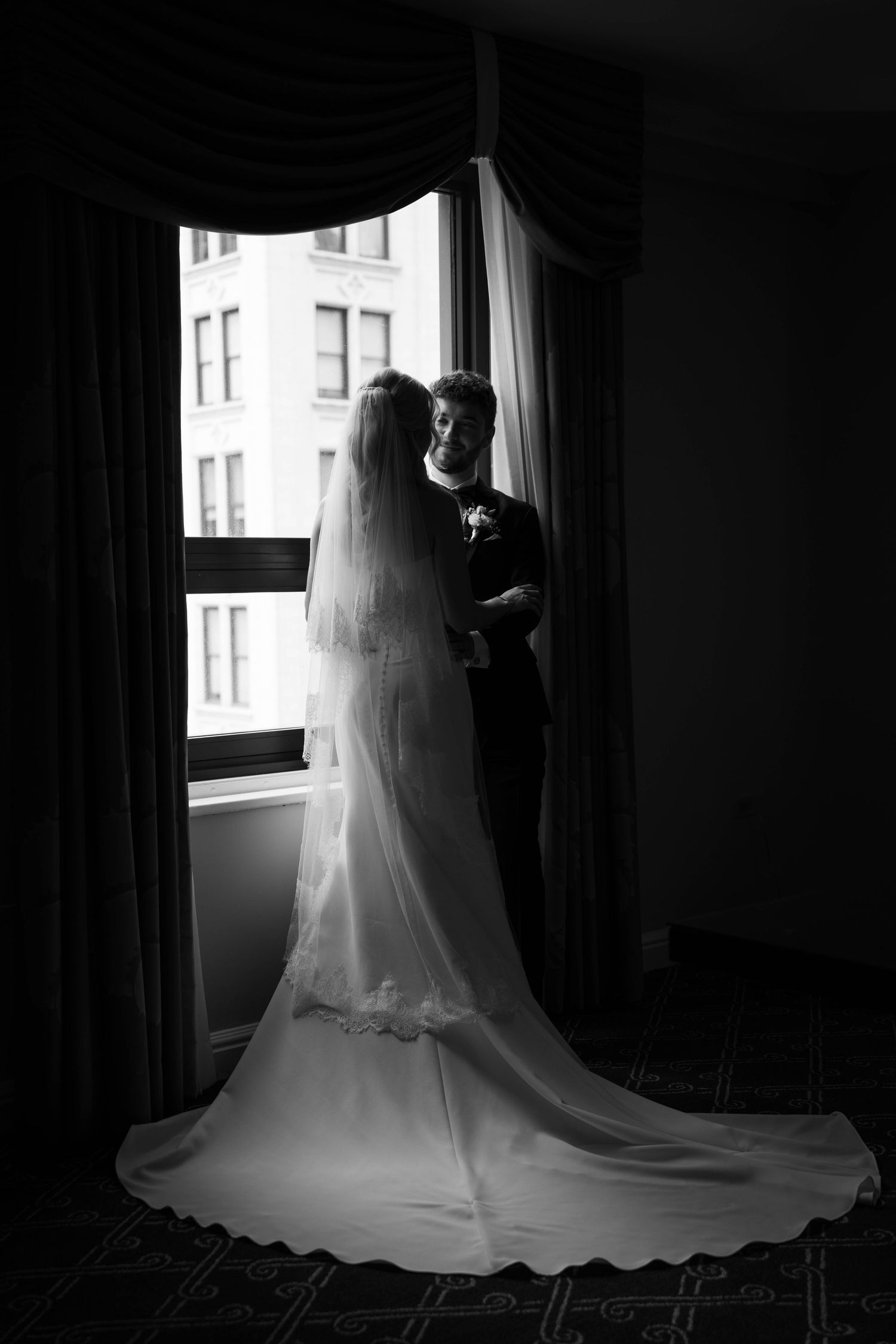 Black and white silhouette of bride and groom embracing by a large window with veil and train flowing.