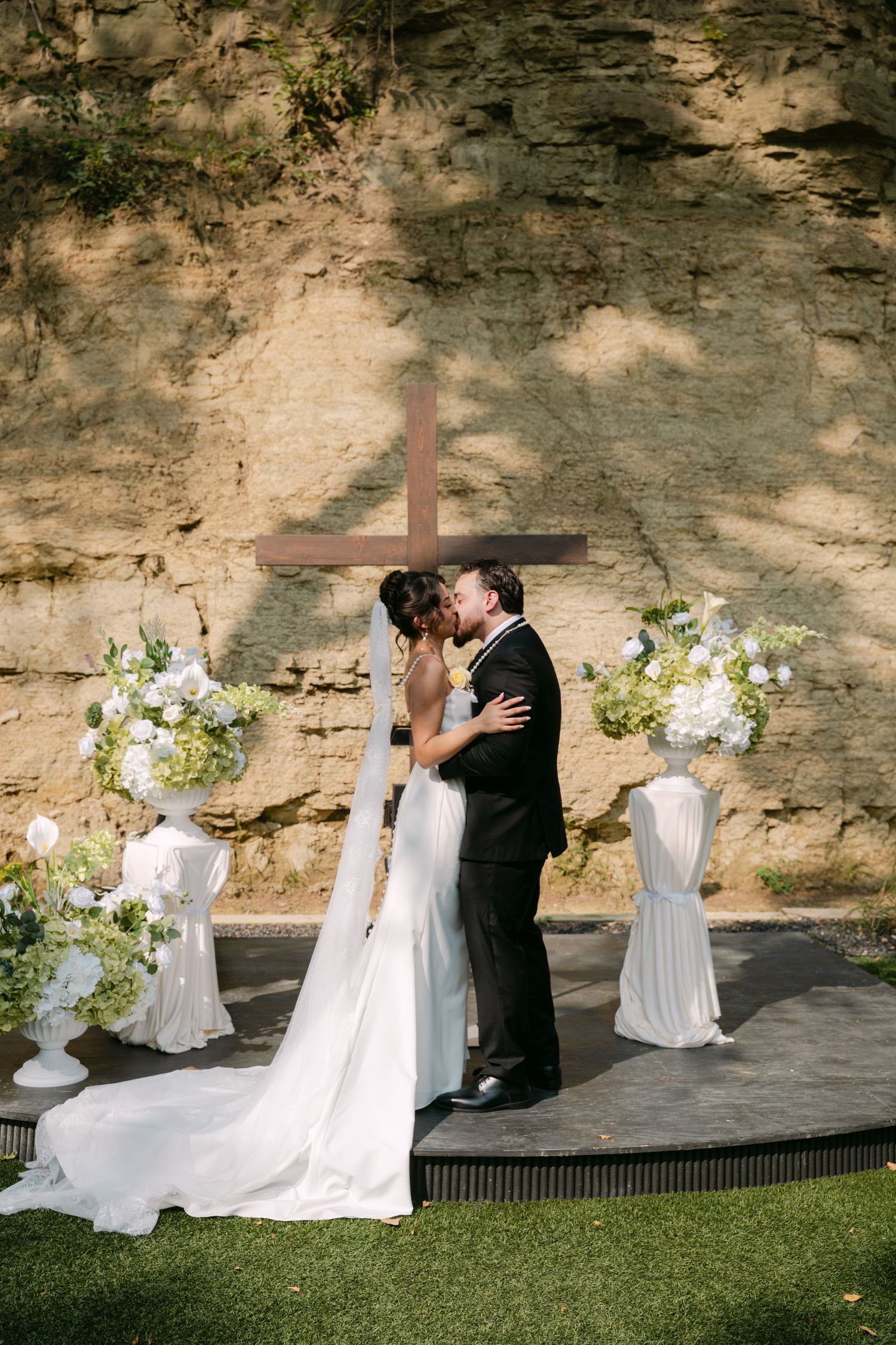 Bride and groom kiss on circular altar with white floral arrangements and wooden cross outdoors.