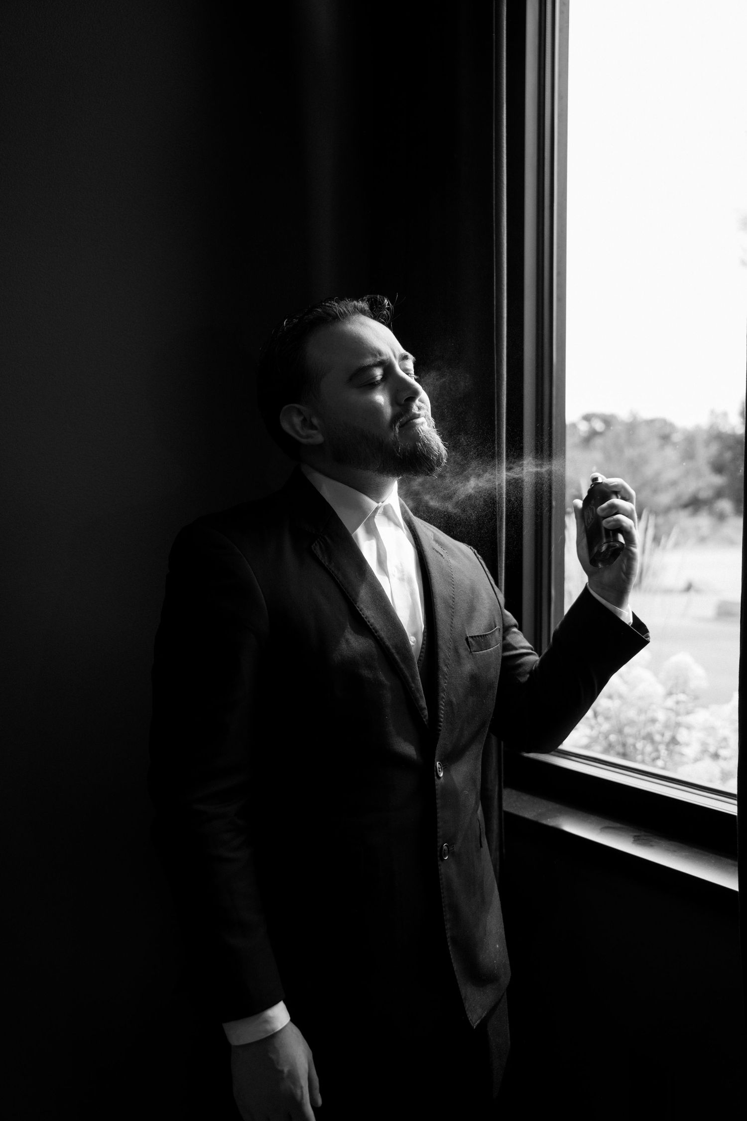 Groom in black suit applying cologne by a window in a moody black and white wedding portrait.