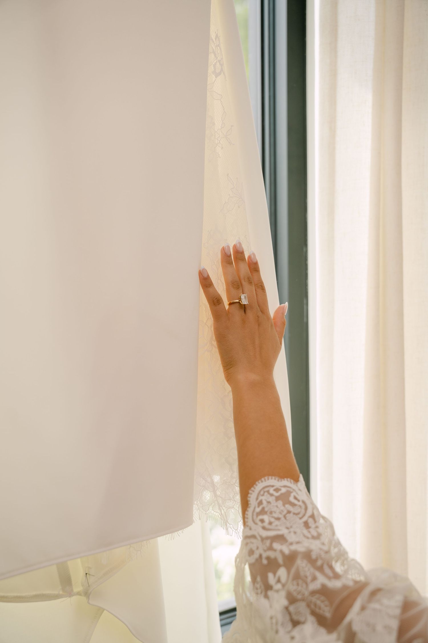 Brides hand with diamond ring gently touching white wedding dress hanging near bright window.