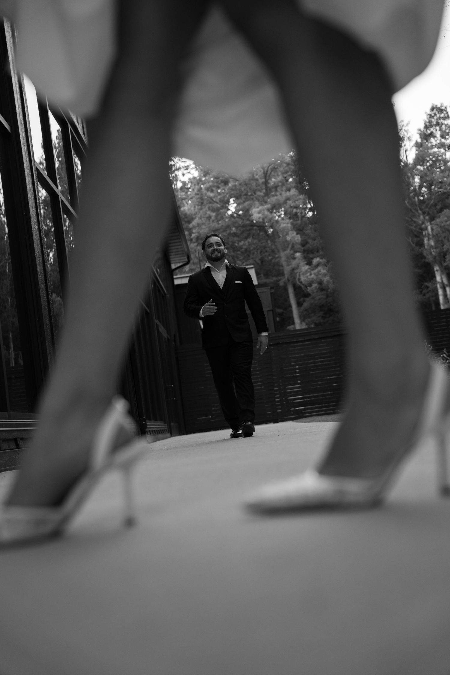 Creative low-angle black and white shot of groom walking toward bride framed by her heels and dress.