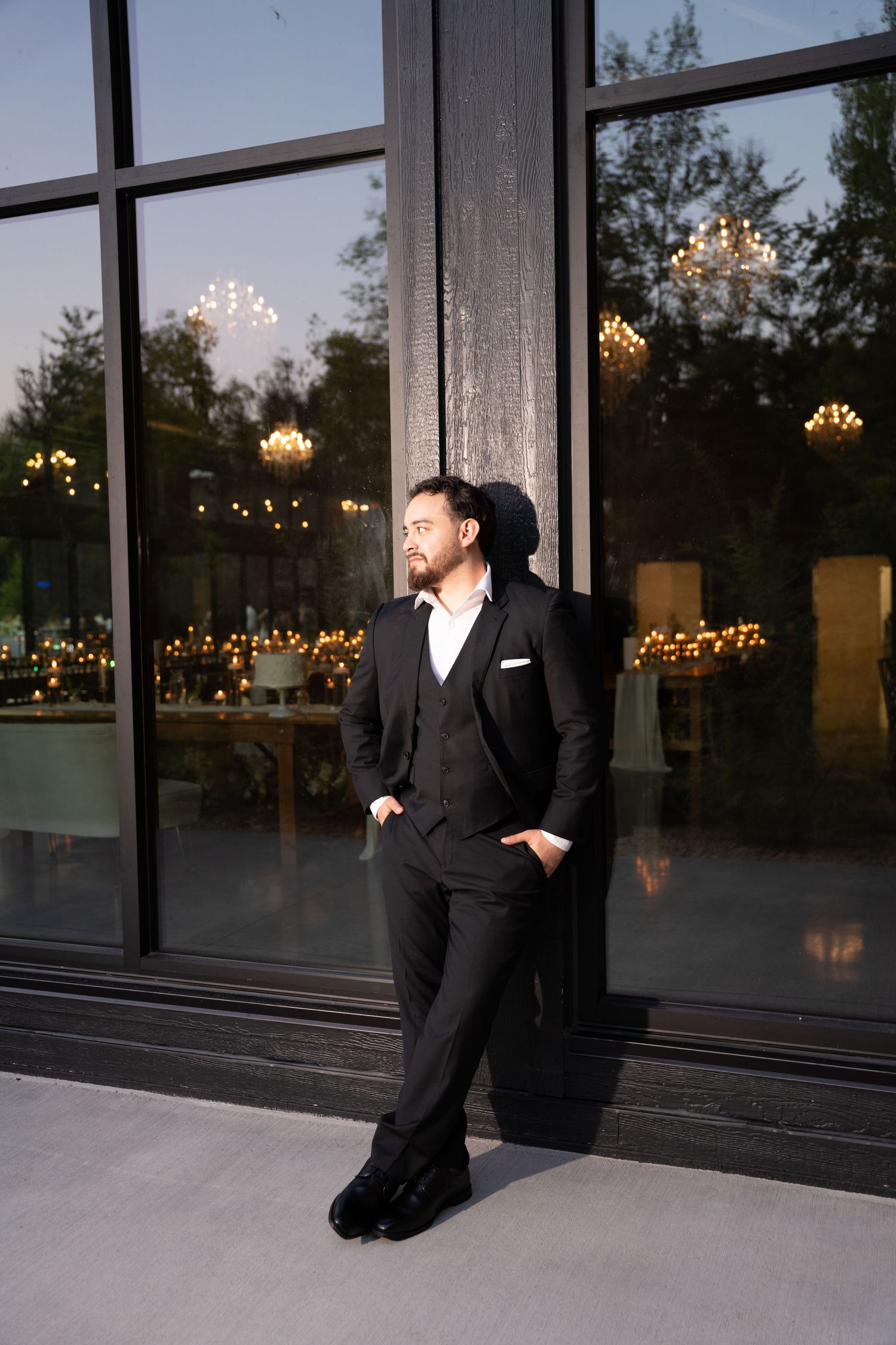 Dapper groom in a black three-piece suit leans against a glass wall reflecting chandelier lights.