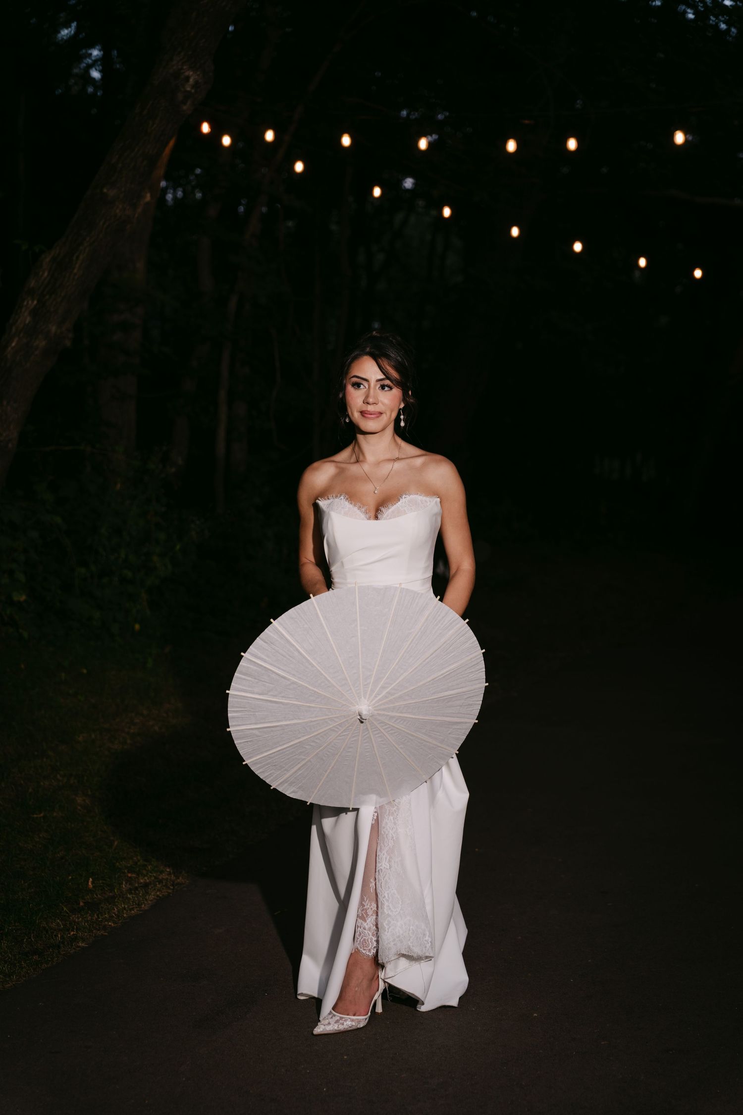 Elegant bride in strapless white gown holding white parasol on tree-lined path with string lights.