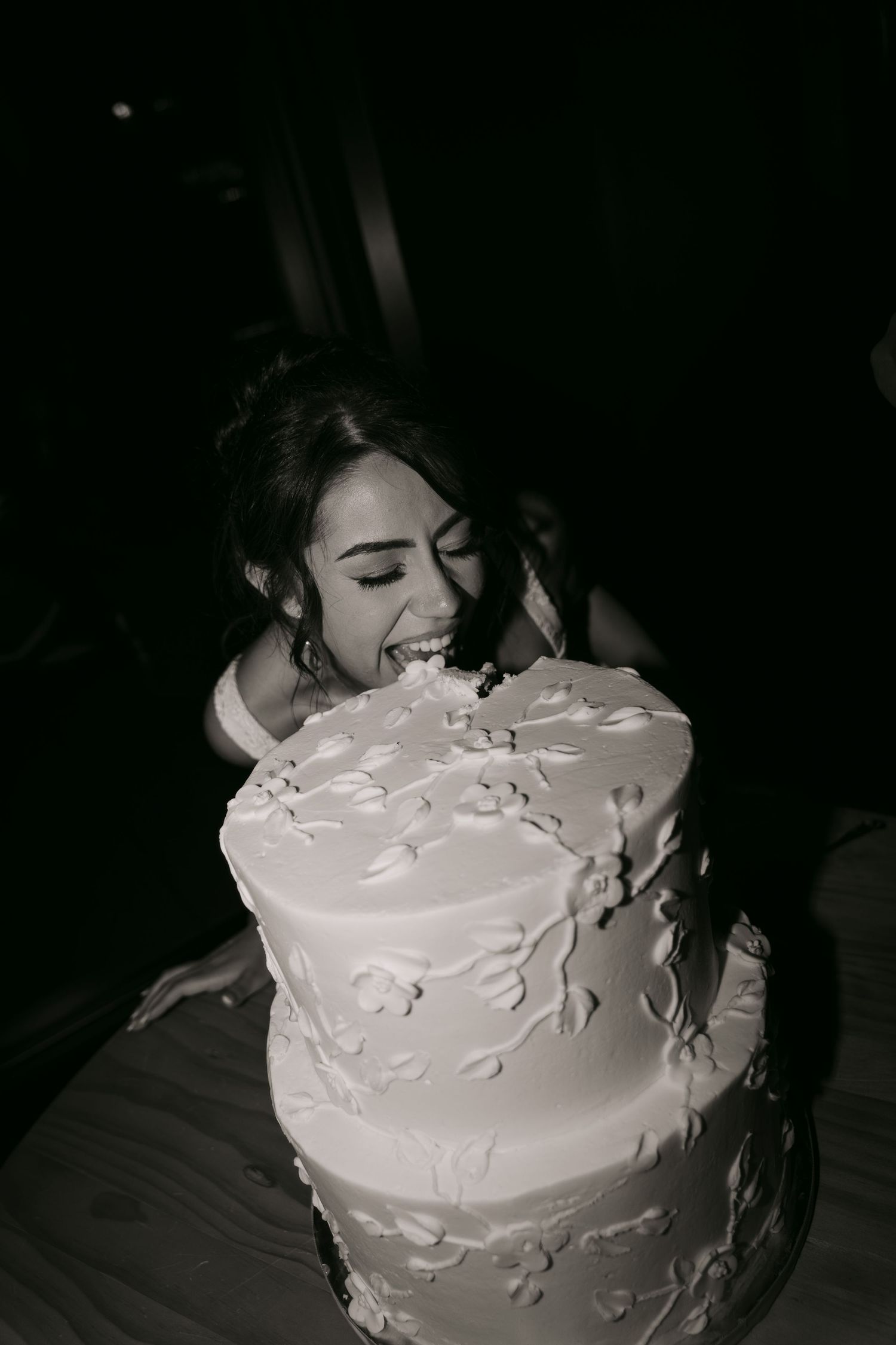 Black and white photo of bride playfully biting into elegant two-tier white floral wedding cake.