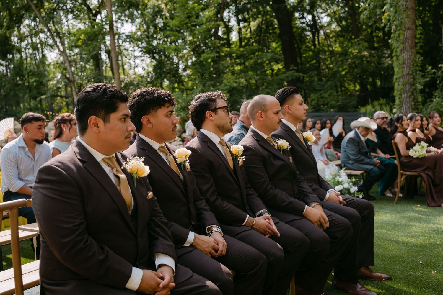Four groomsmen in dark suits with yellow boutonnieres seated at outdoor wedding ceremony.