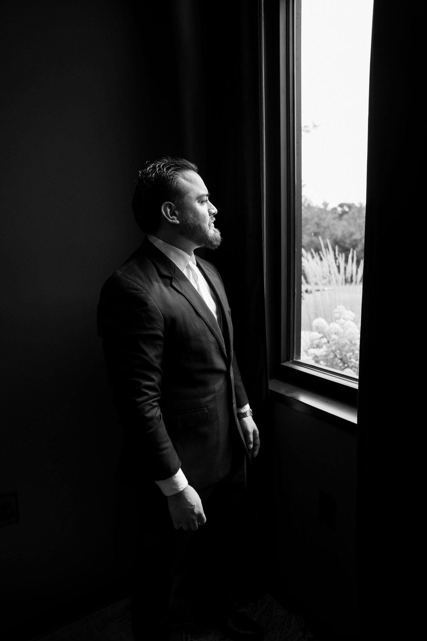 Black and white portrait of groom in dark suit gazing out a large window with natural light.