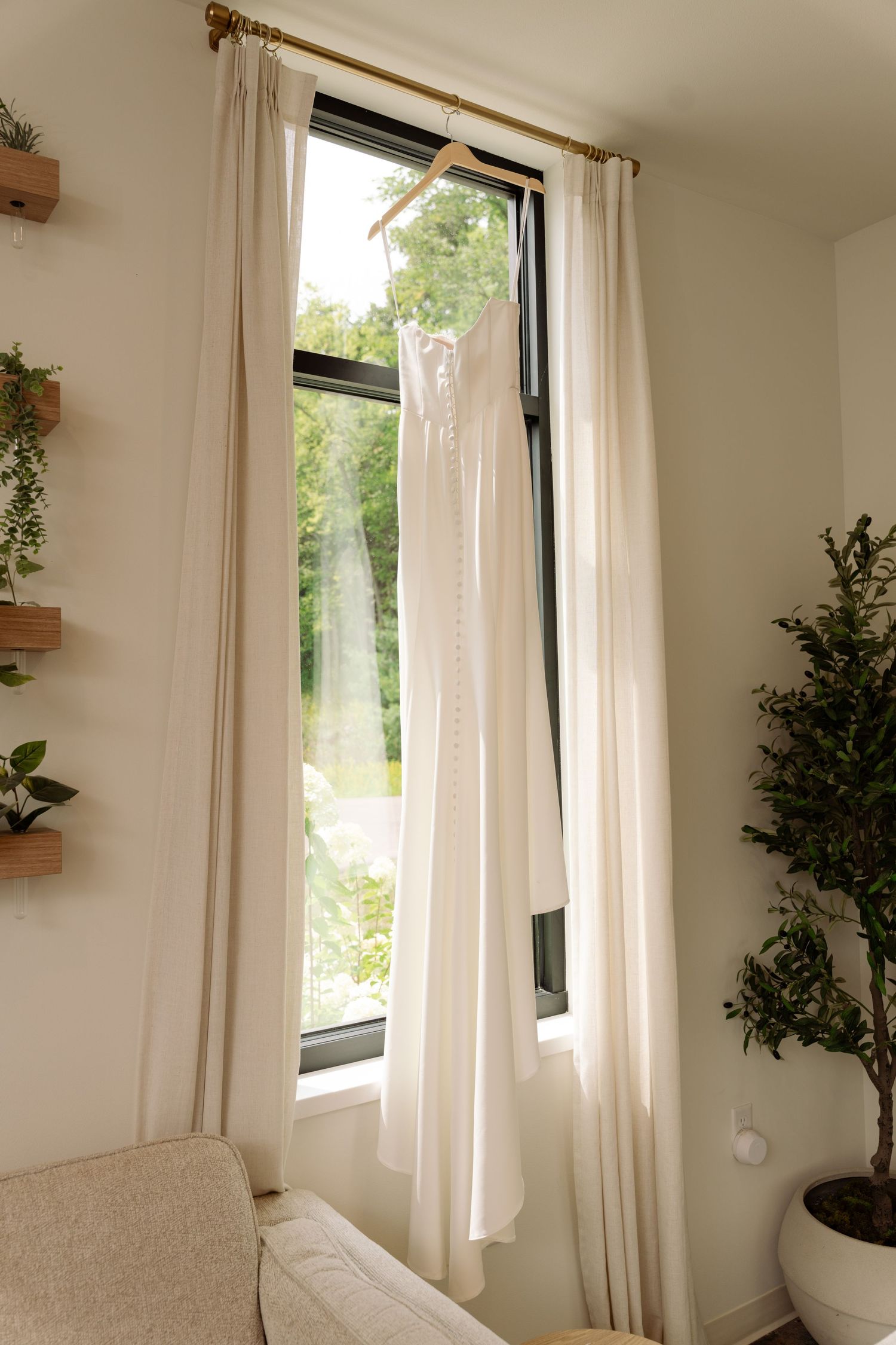 White wedding dress hanging on window in sunlit room with curtains and green plants nearby.