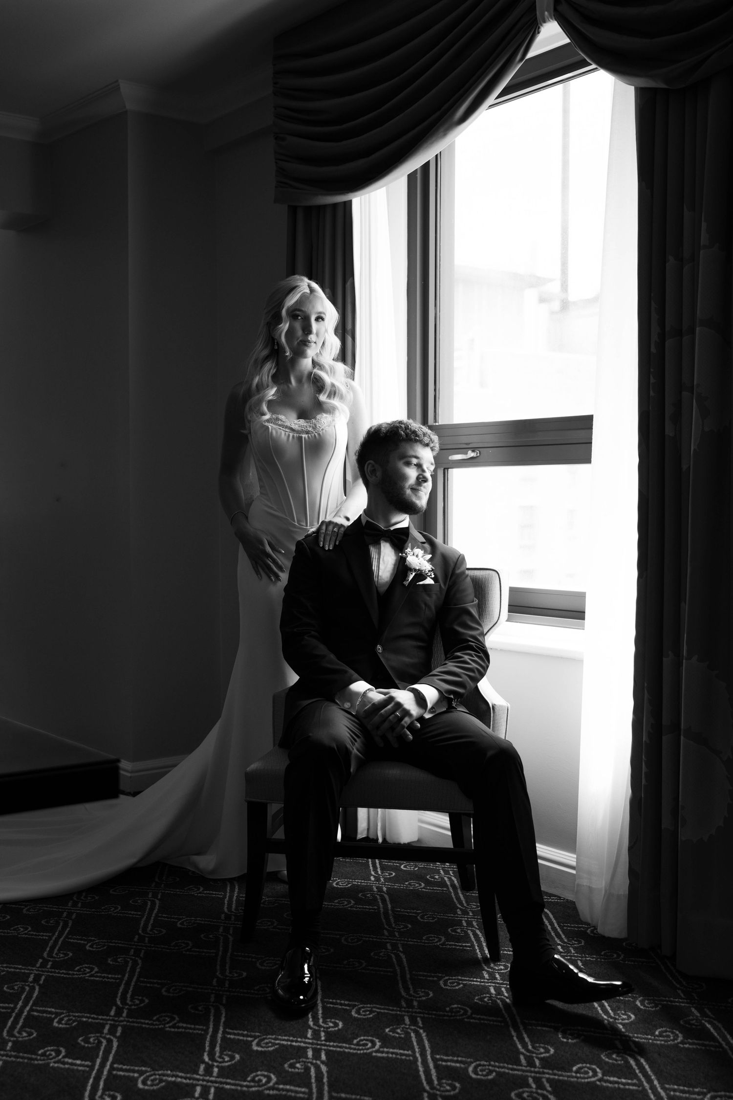 Black and white wedding photo of bride standing behind seated groom near window in elegant hotel room.