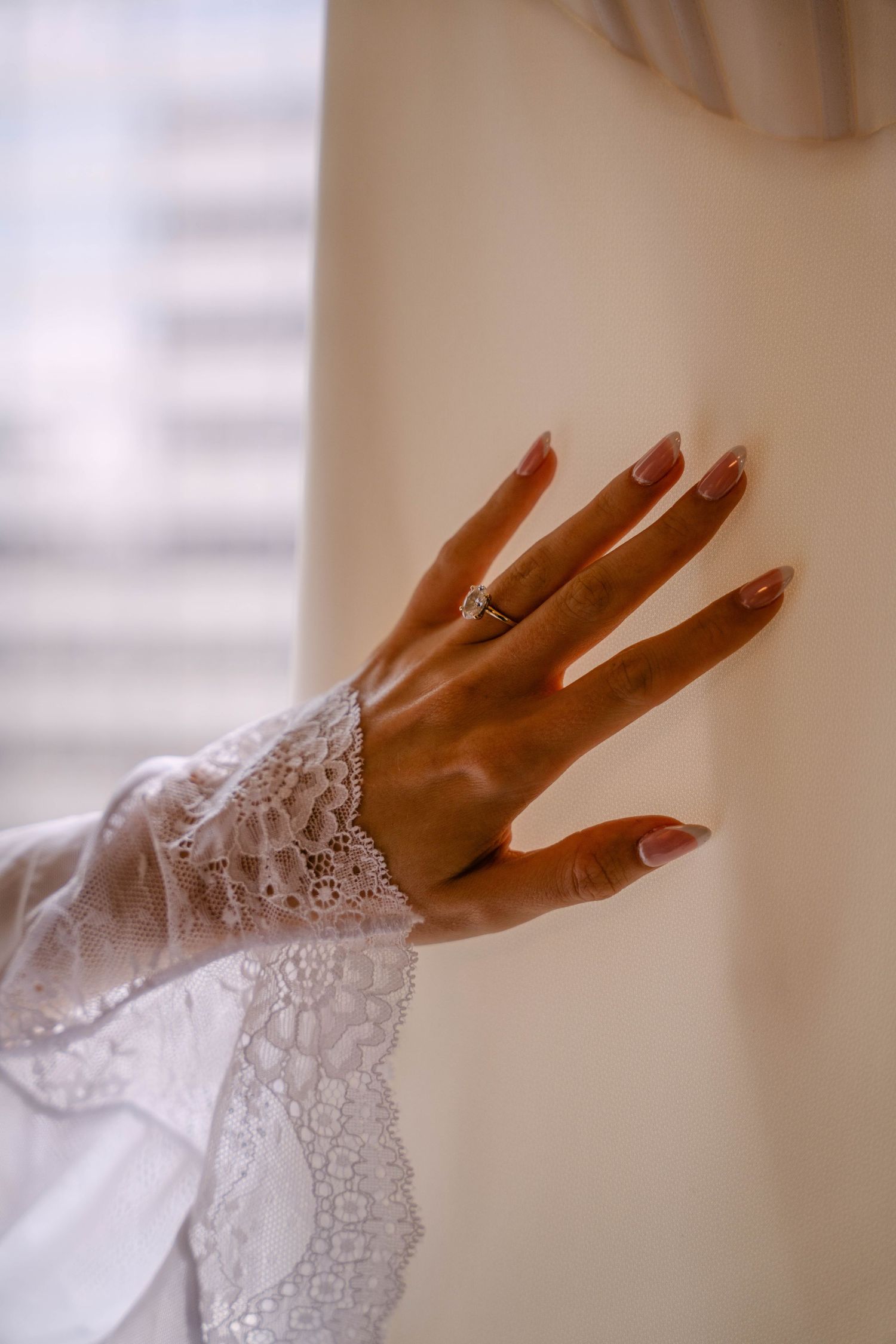 Bride showcasing elegant engagement ring with French manicure, wearing white lace wedding dress sleeve.