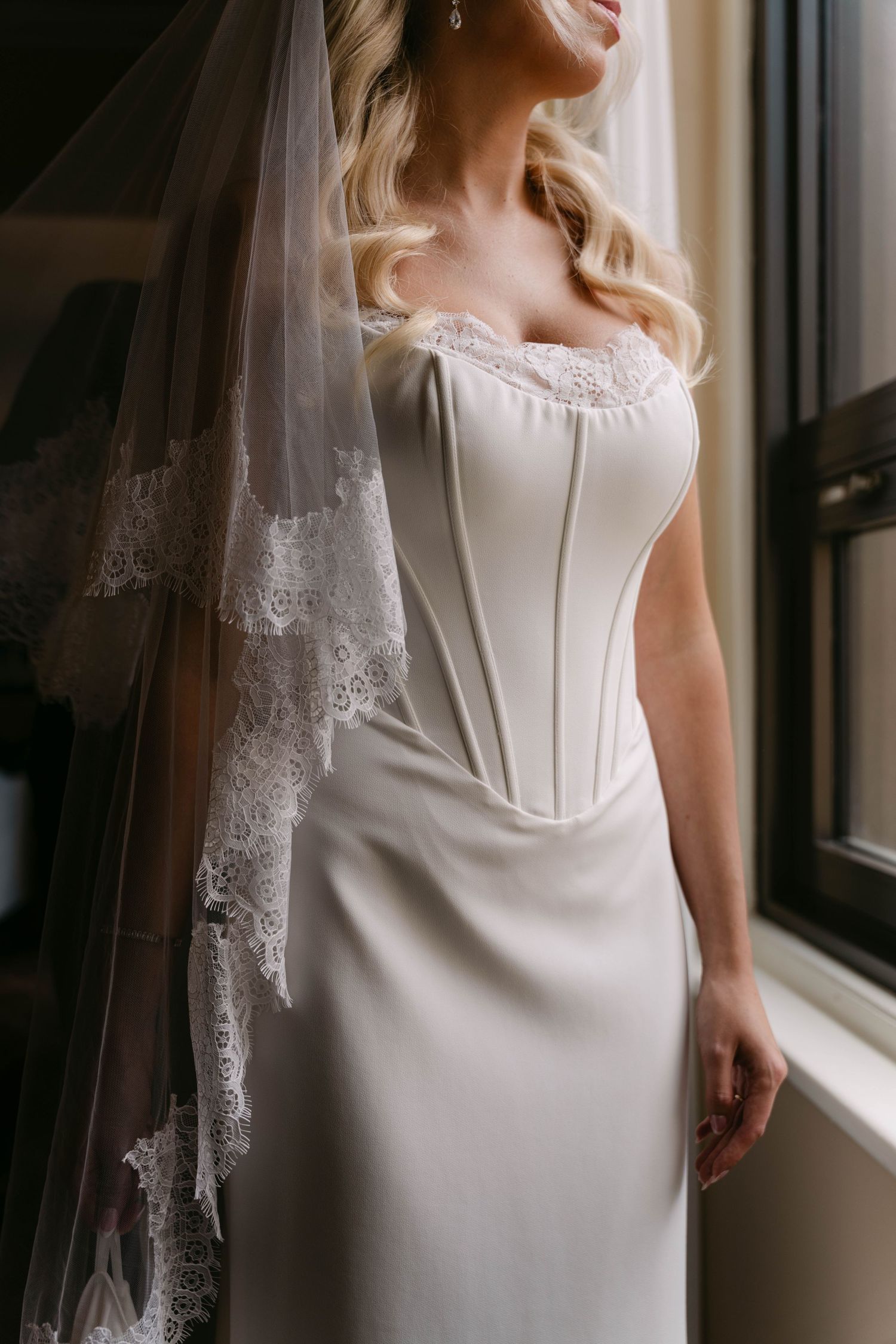 Elegant bride in a white corset wedding gown with lace veil by a window.