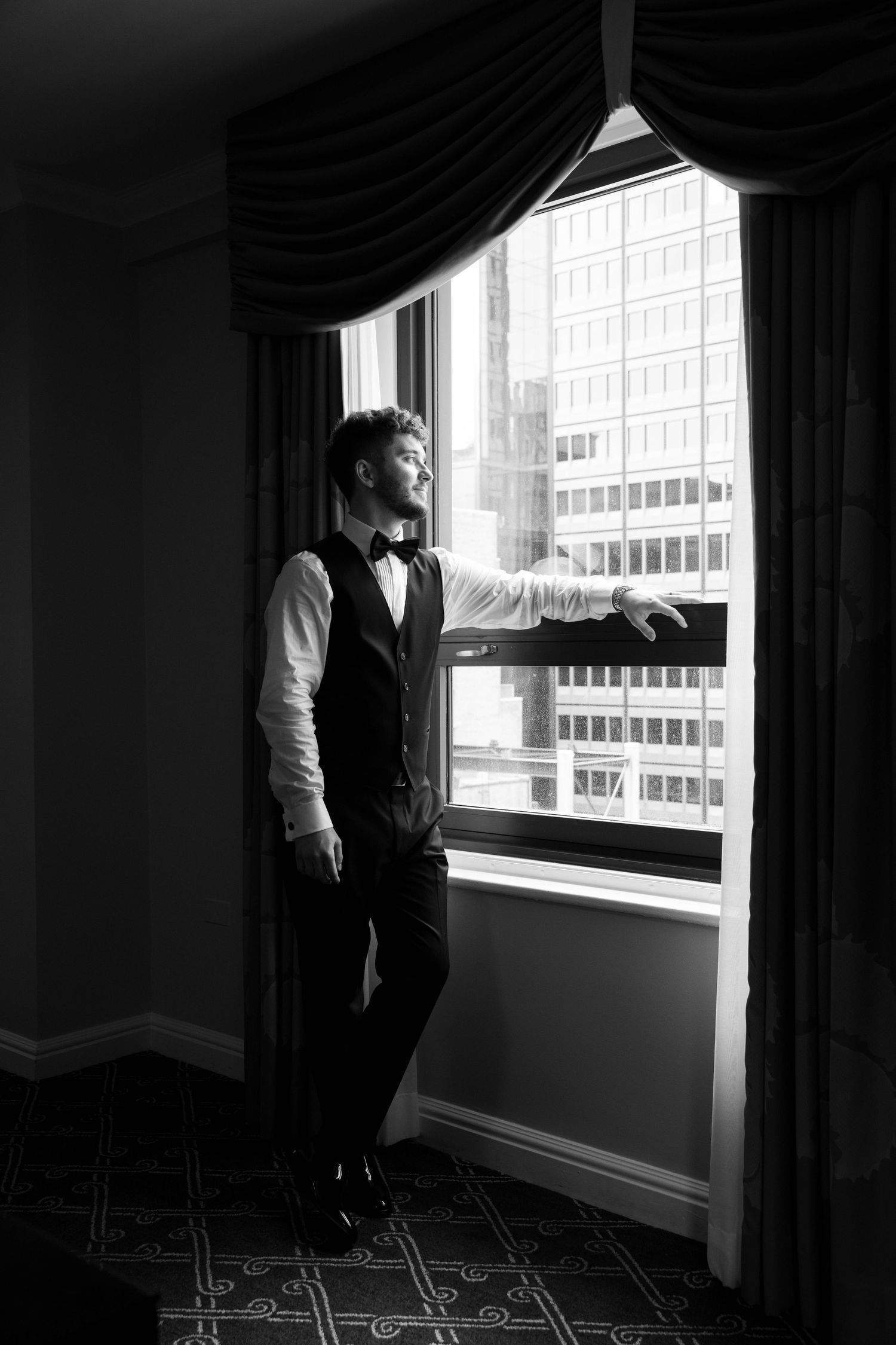 Groom in vest and bow tie gazes out hotel window at city skyline in elegant black and white wedding portrait.