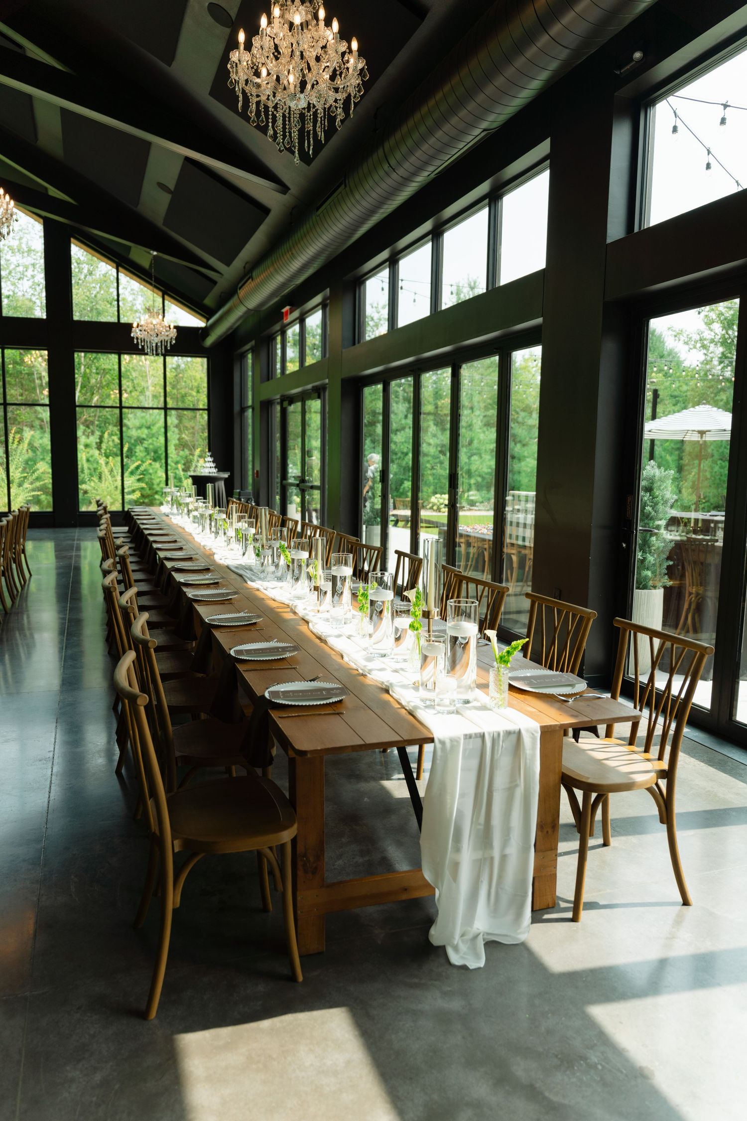Elegant wedding reception hall with long wooden tables, chandeliers, and floor-to-ceiling windows.