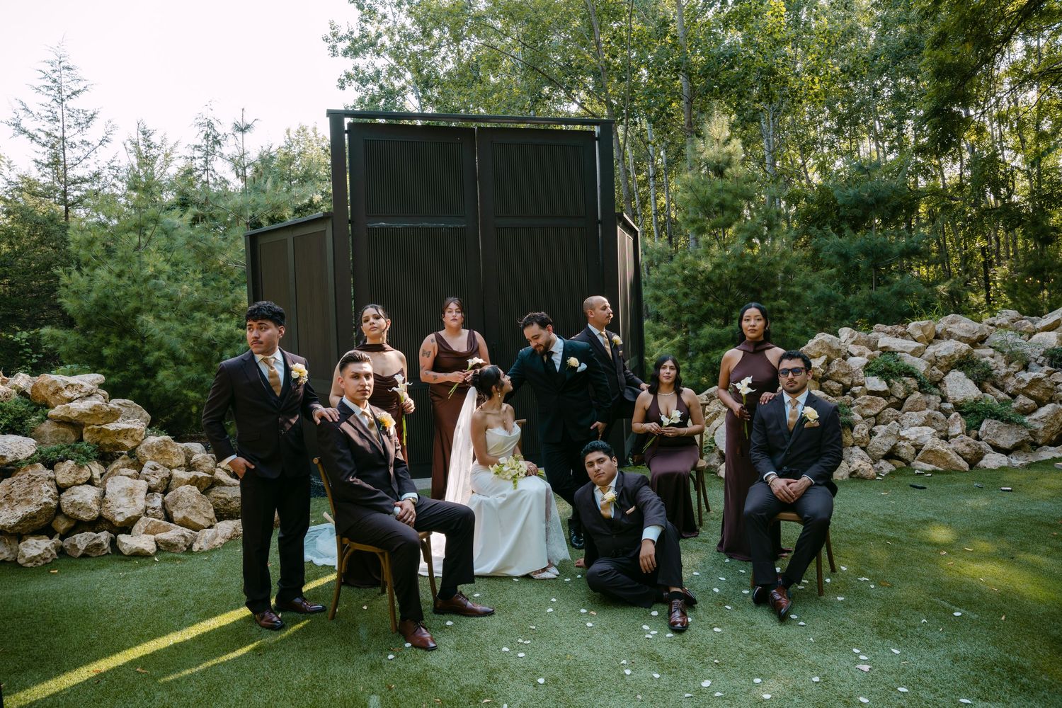 Wedding party group photo on green lawn with stone wall background and dark chapel structure.