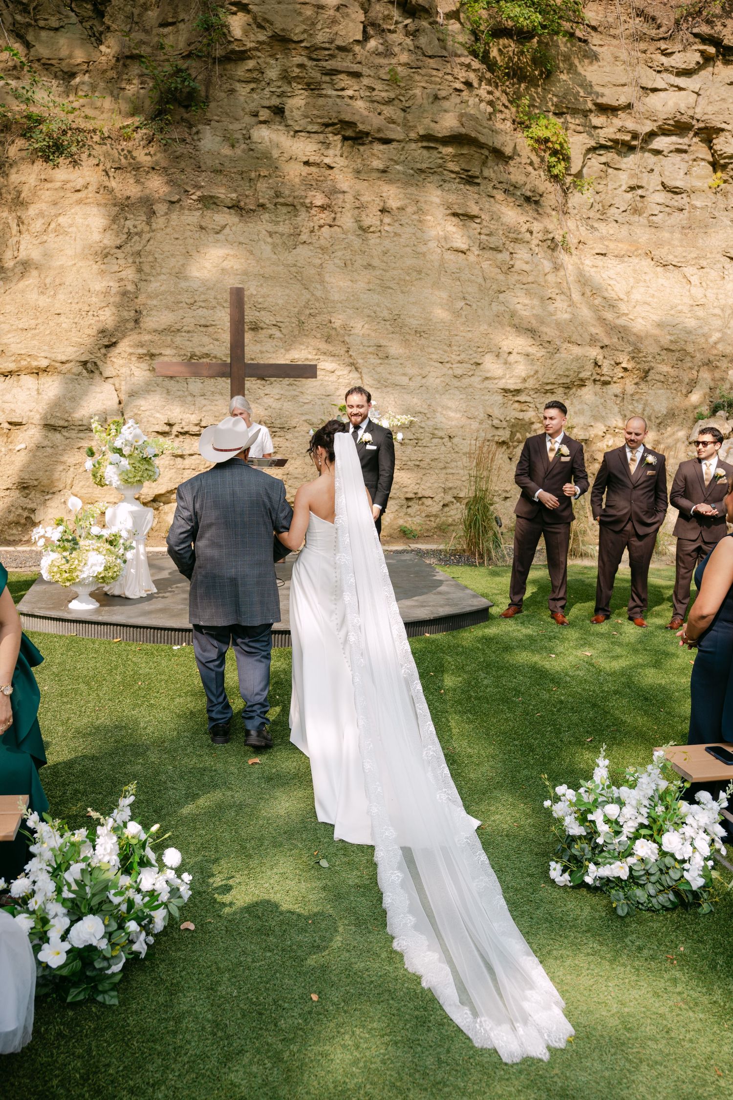 Aerial view of bride with cathedral veil approaching smiling groom at outdoor wedding altar with cross.