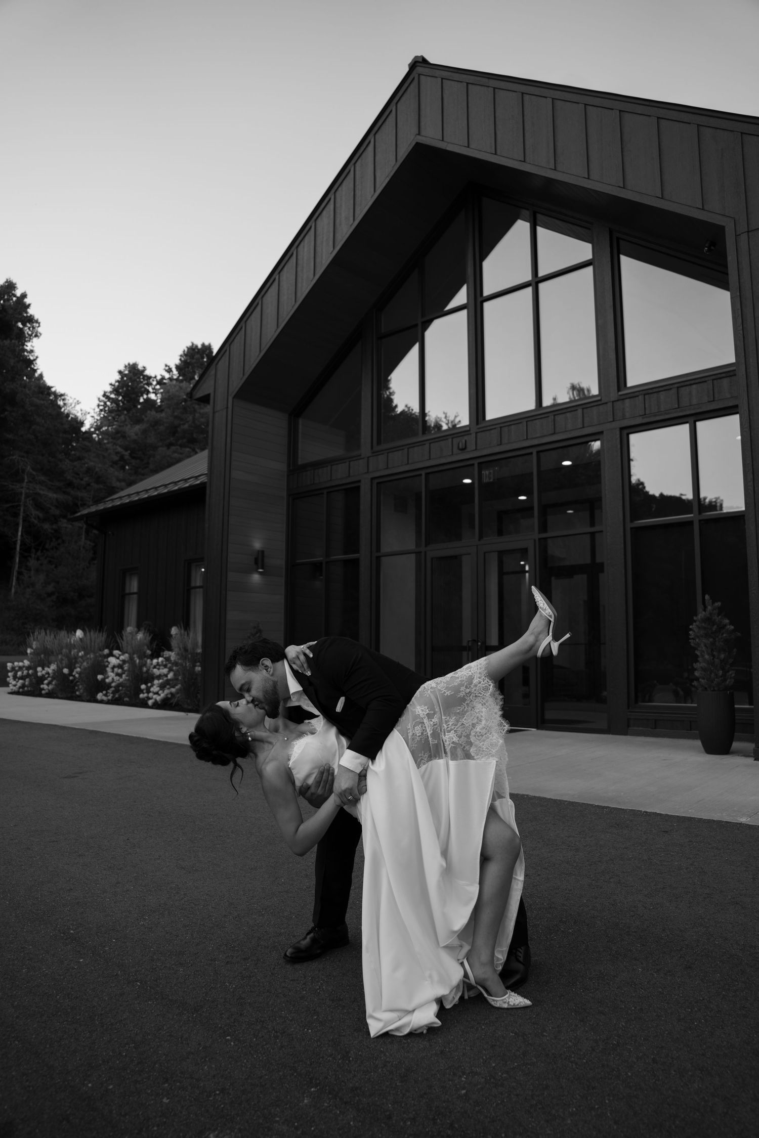 Bride and groom share a romantic dip kiss outside a modern wedding venue at dusk in black and white.