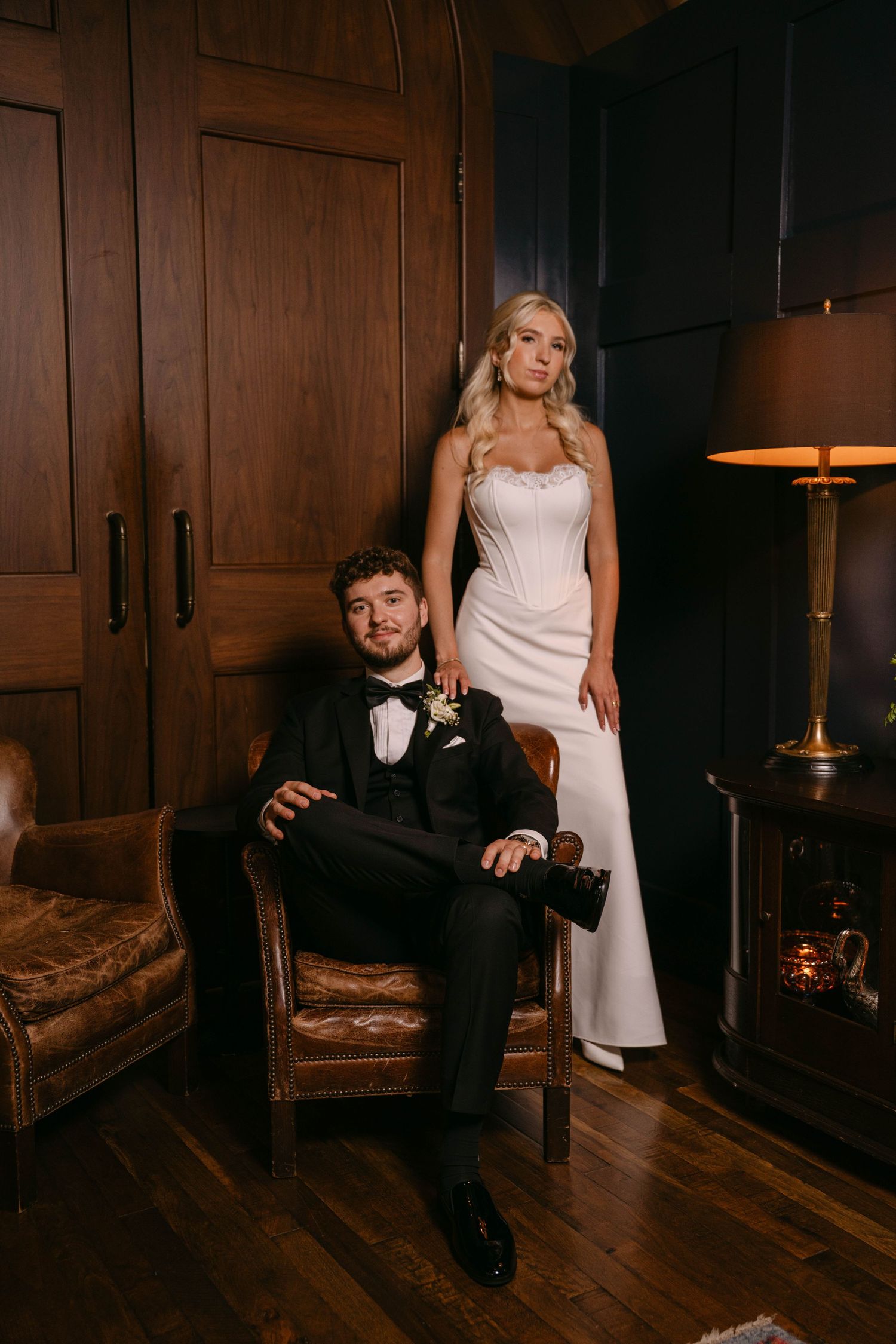 Bride standing beside seated groom in a dark wood-paneled room with vintage leather chair and lamp.