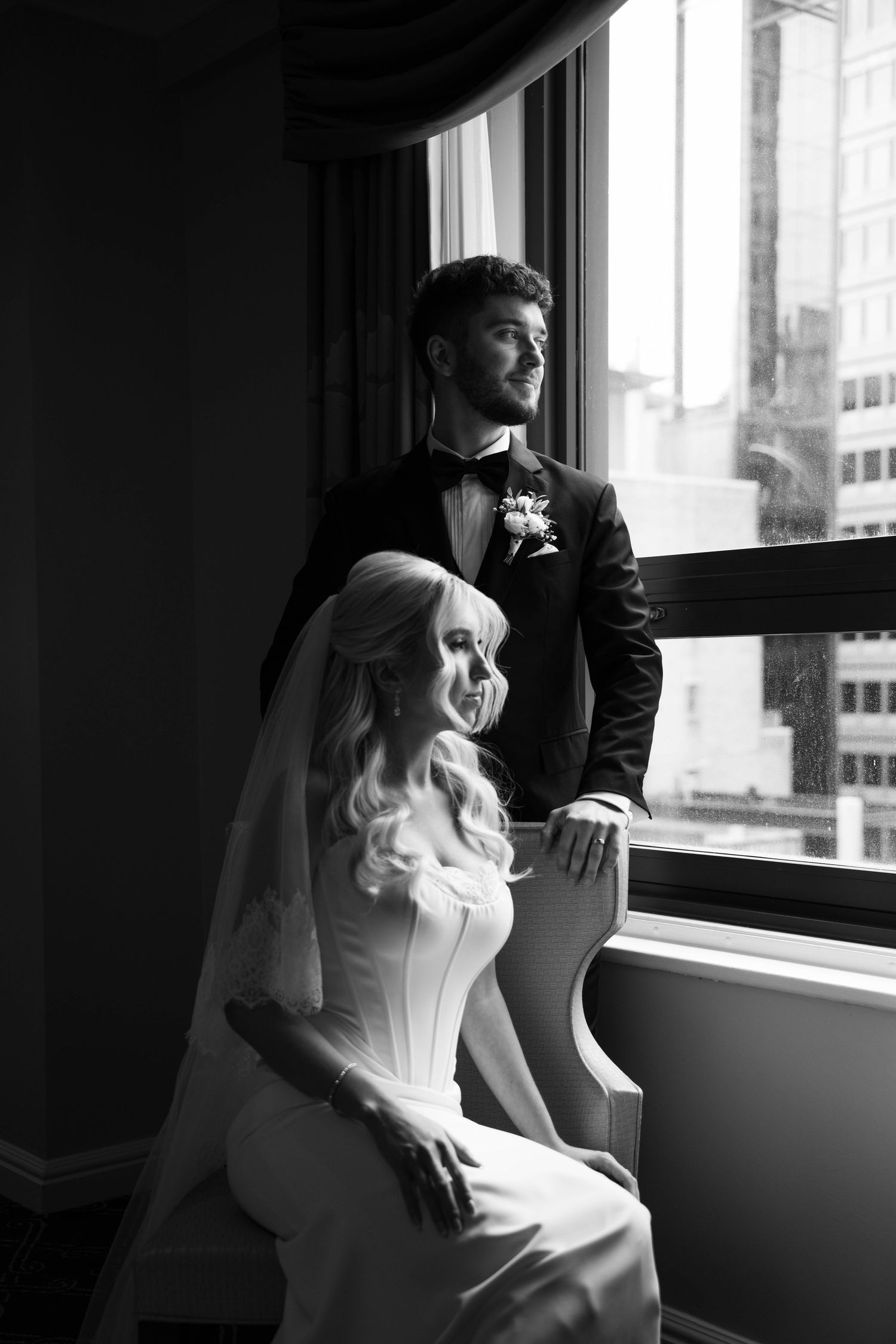 Black and white photo of bride and groom posing by a large hotel window.