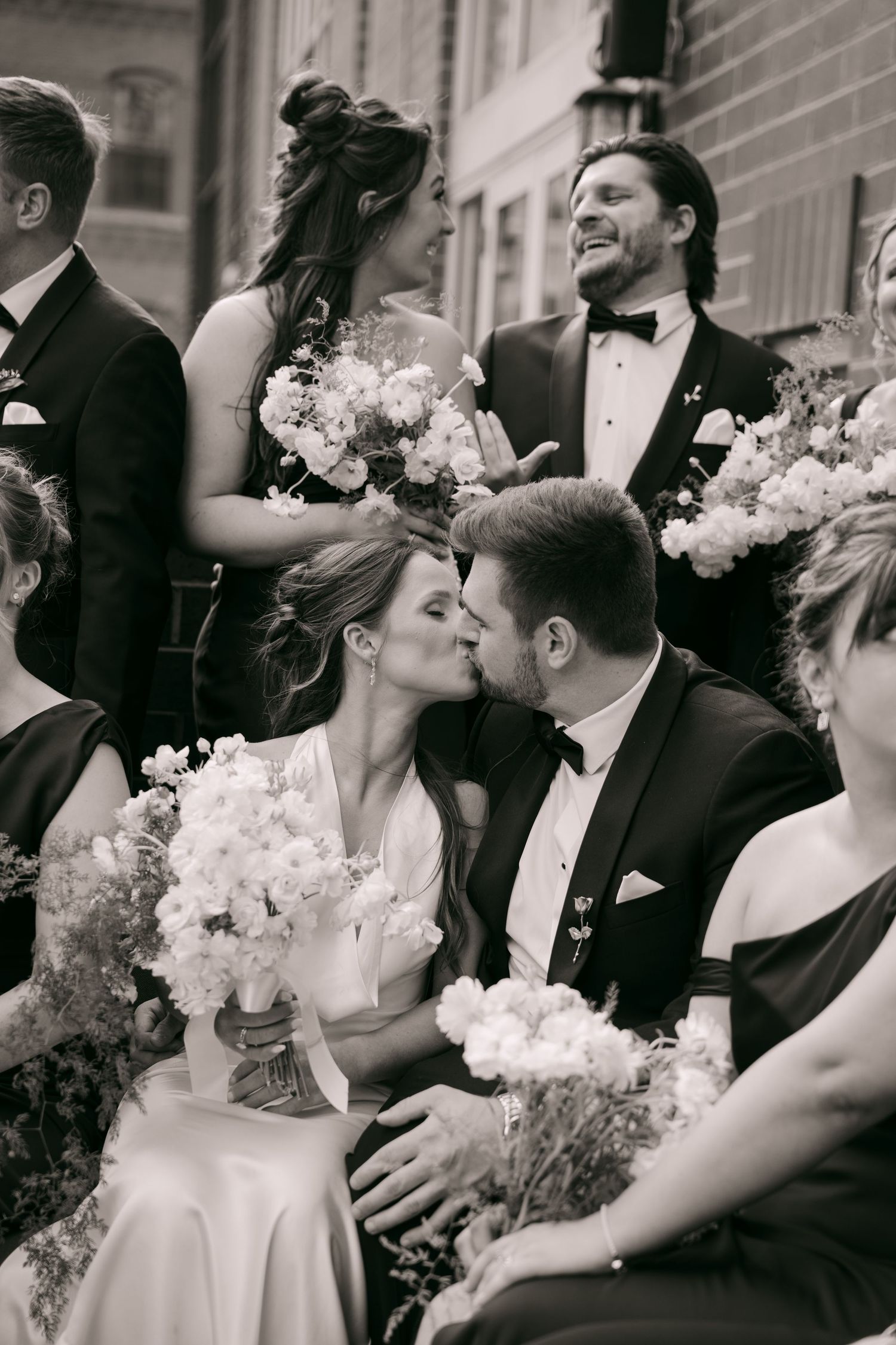 Black and white photo of bride and groom sharing a kiss surrounded by wedding party holding floral bouquets.