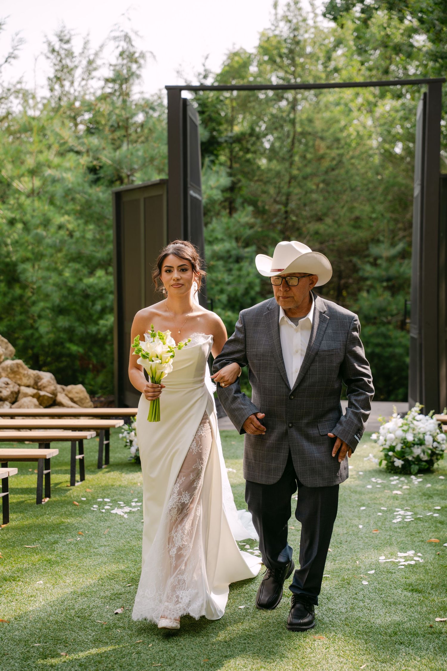 Bride in strapless white gown holding calla lily bouquet walks down aisle with man in cowboy hat.