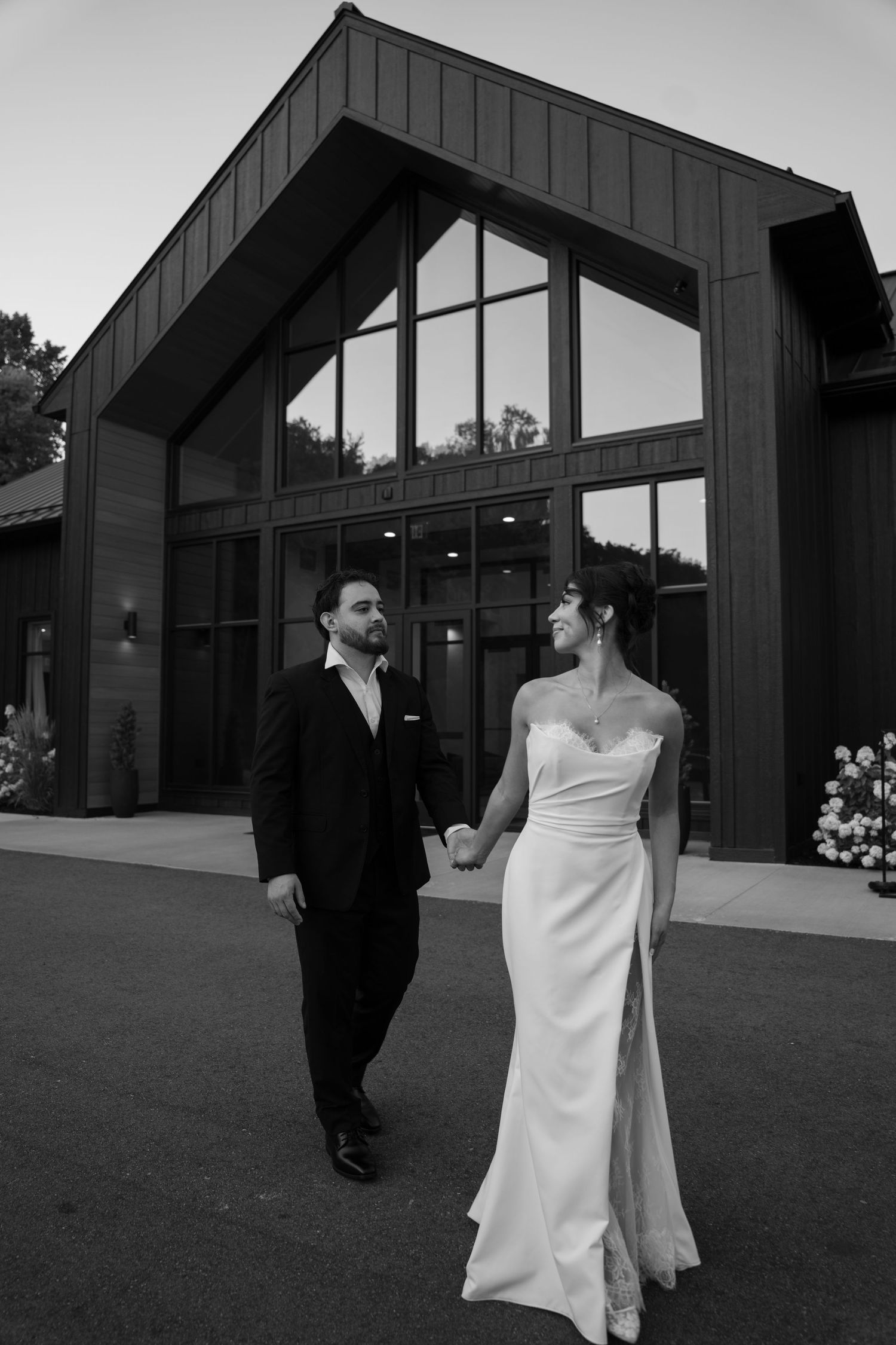 Bride and groom walk hand in hand outside a modern wedding venue at dusk.