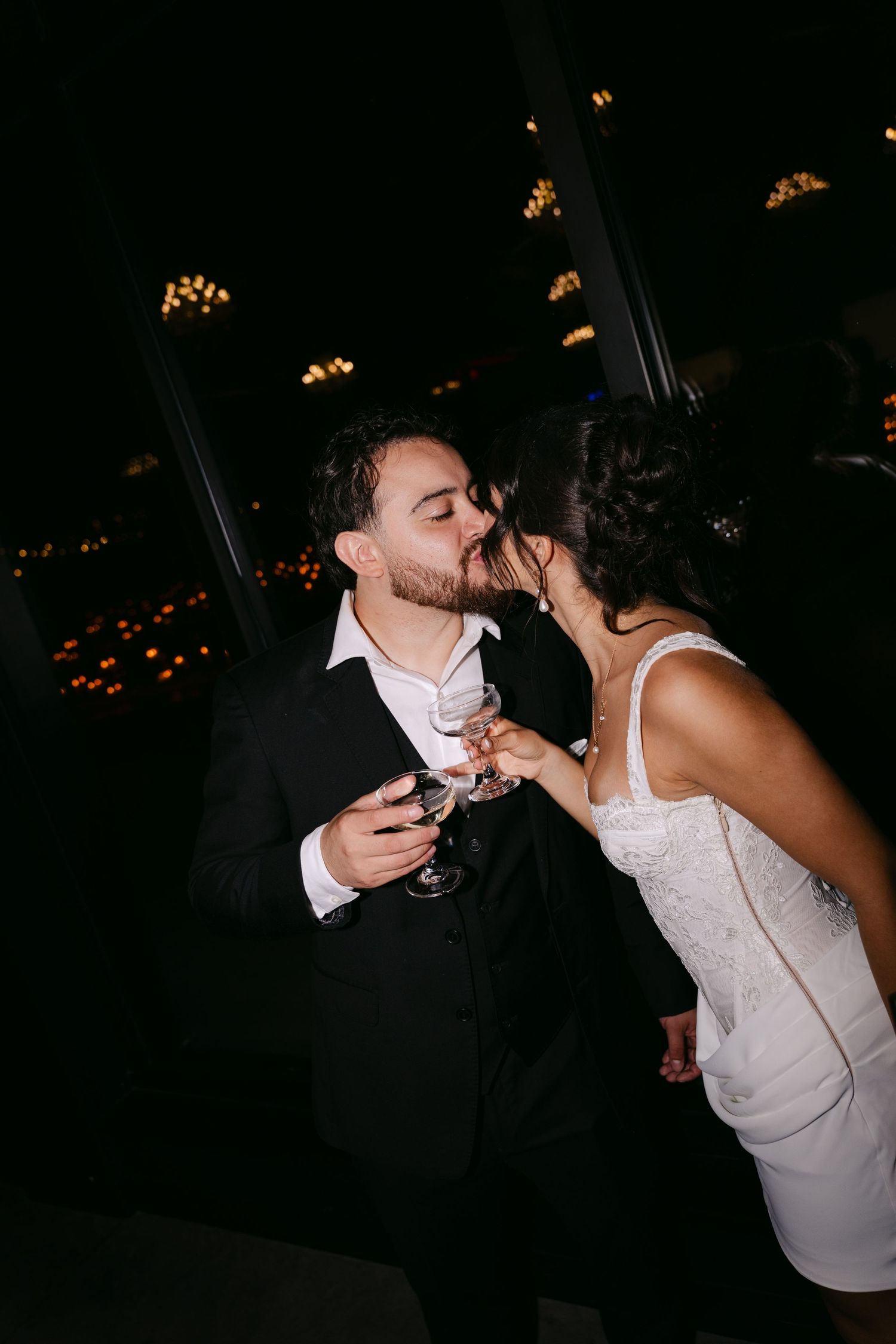 Bride and groom sharing a romantic kiss while holding champagne glasses at dark wedding reception.