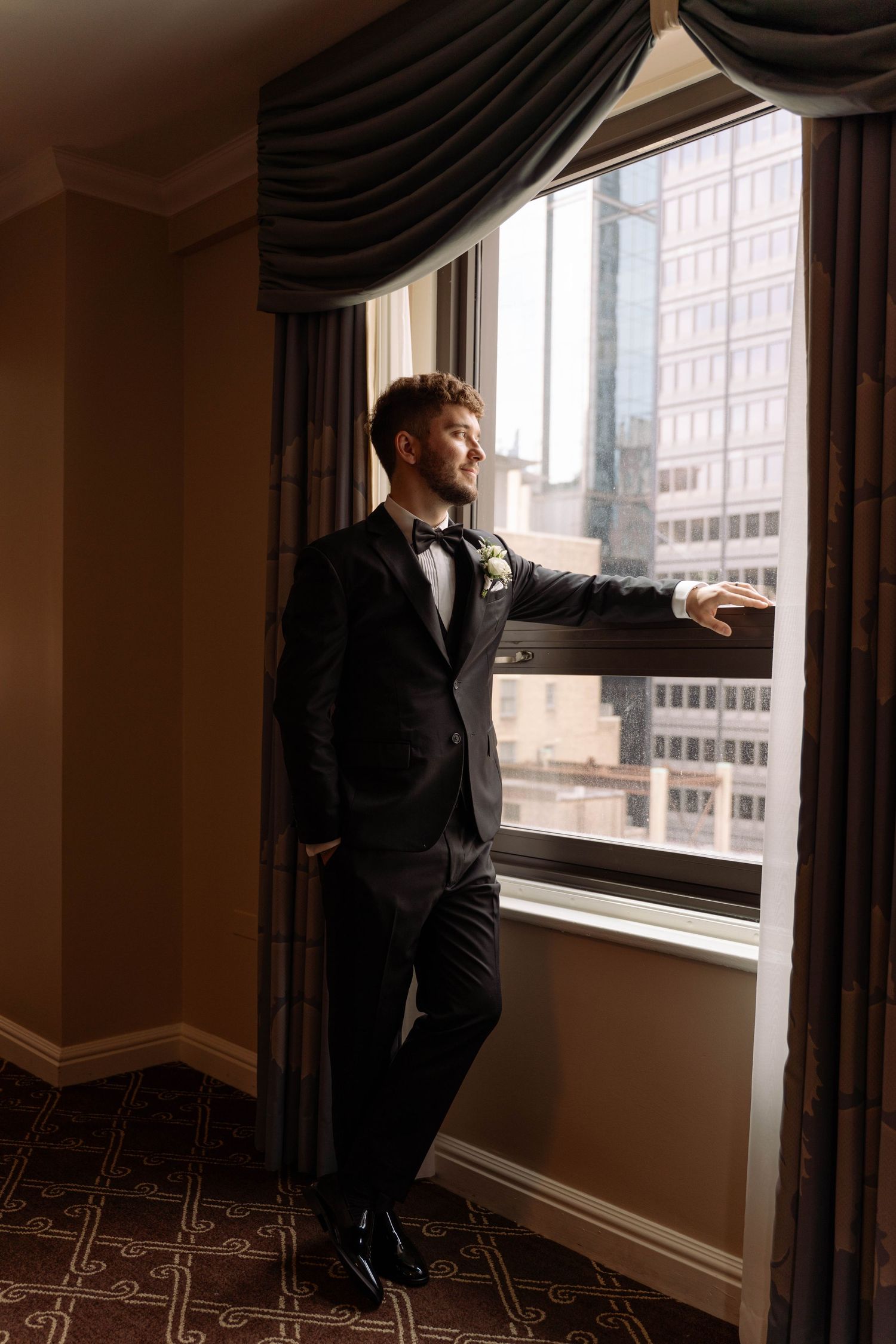 Groom in black tuxedo standing by hotel window overlooking city buildings.