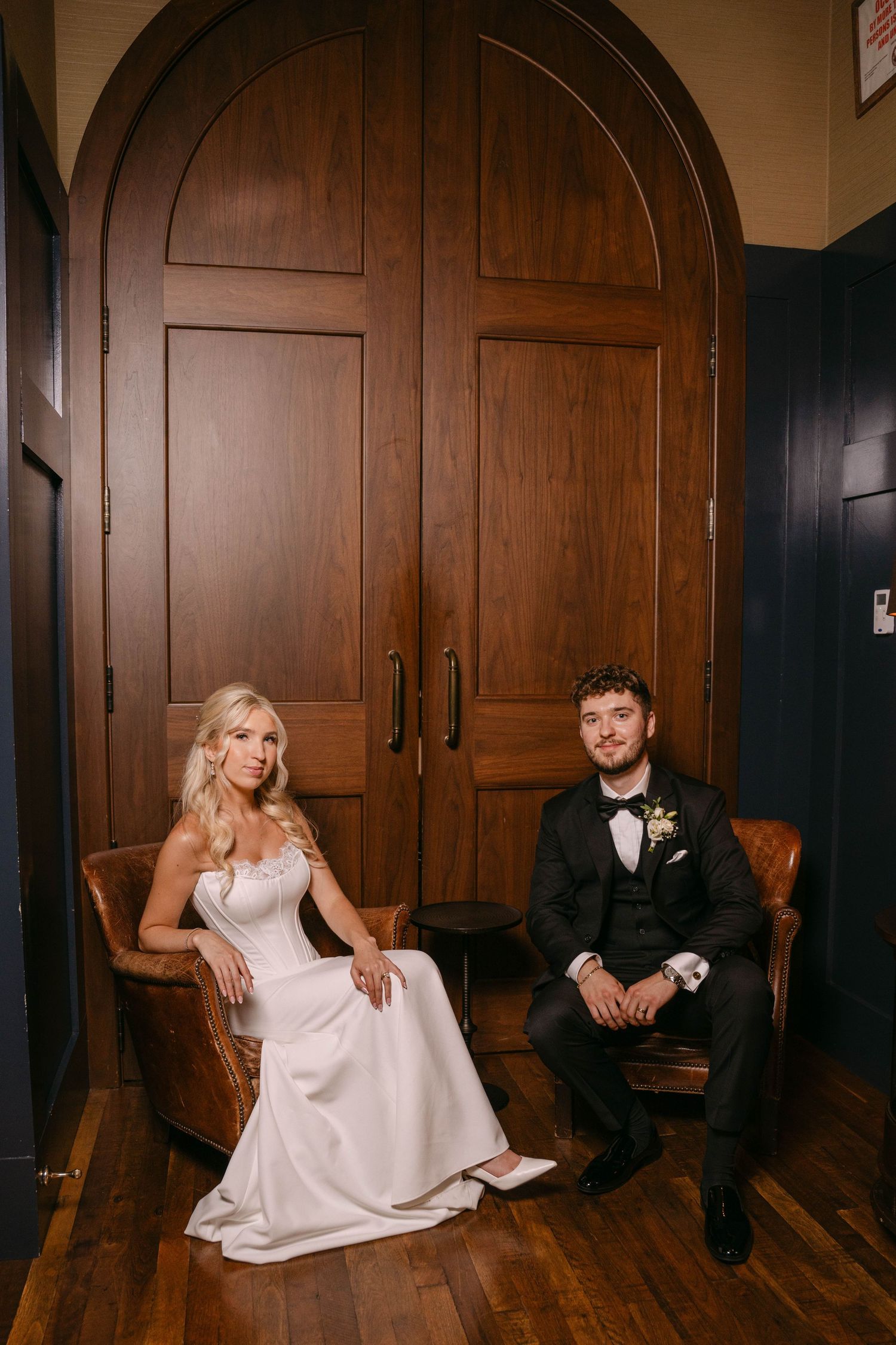 Bride and groom seated in leather chairs before arched wooden doors at venue.
