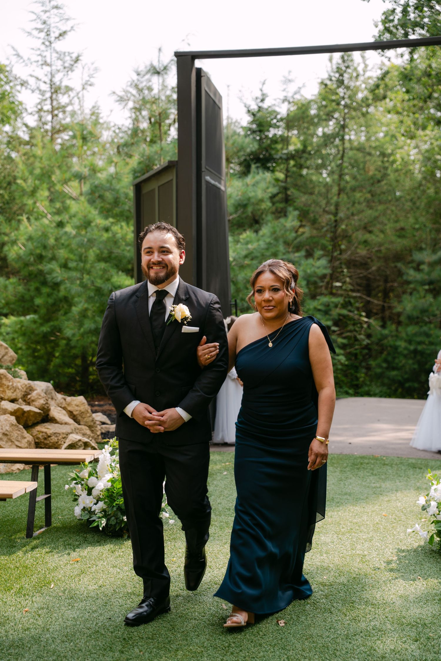 Groom in black suit with boutonniere walks down aisle with woman in teal gown at outdoor wedding ceremony.