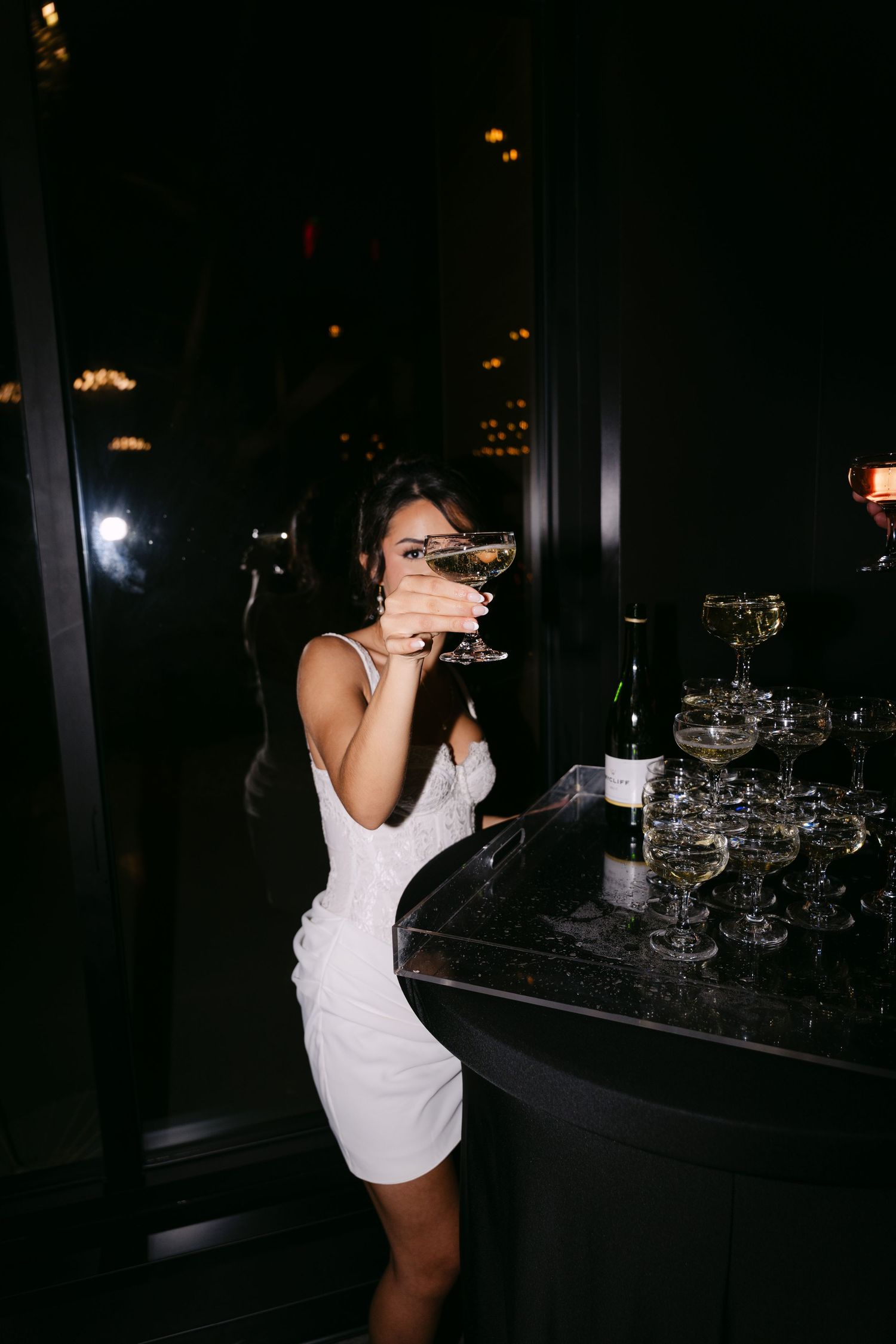 Bride in white mini dress holding champagne coupe glass at elegant dark wedding reception bar.