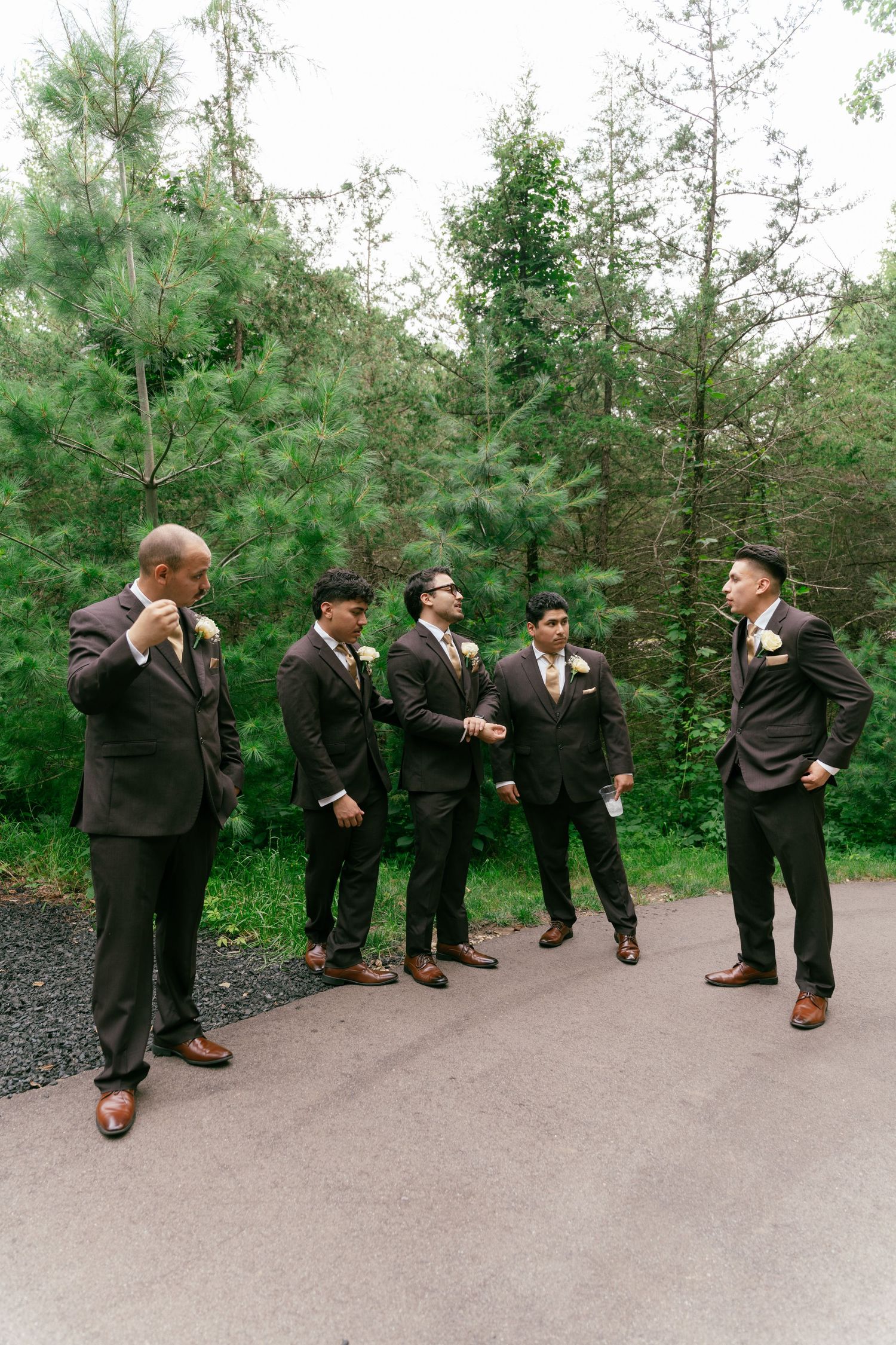 Groom and groomsmen in dark suits with boutonnieres gathered on pathway surrounded by pine trees.