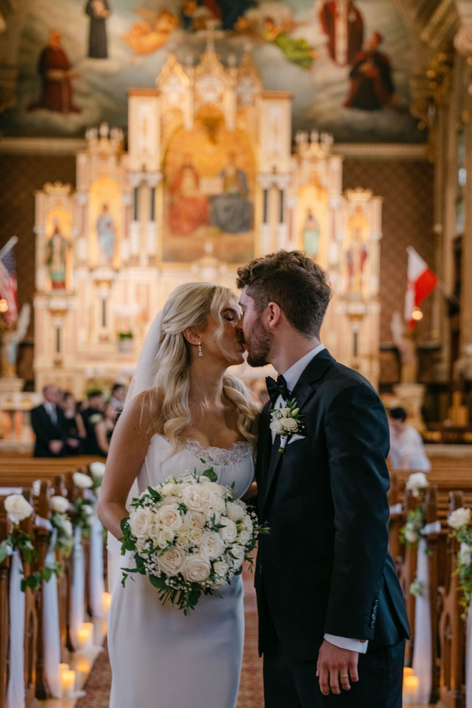 Bride and groom sharing first kiss at ornate church altar, bride holding white rose bouquet.