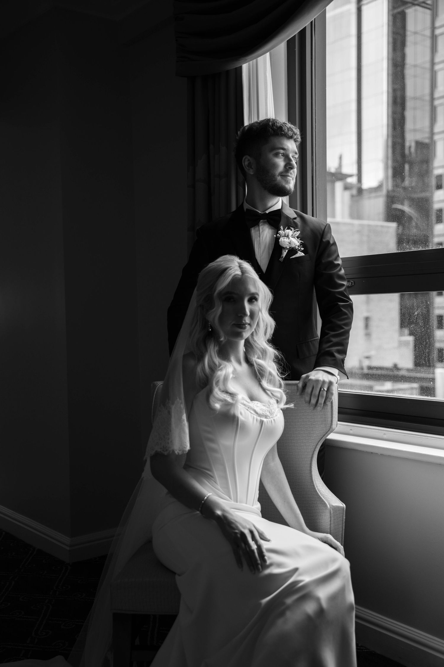 Black and white wedding photo of bride in white gown seated by window with groom in tuxedo standing beside her.