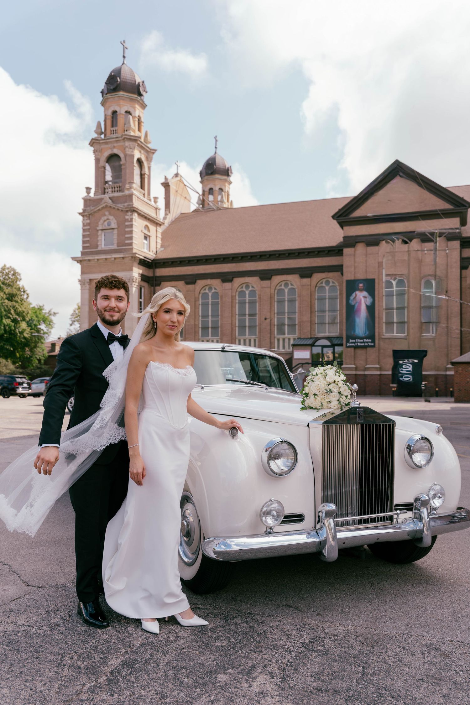 Bride and groom posing beside vintage white Rolls-Royce in front of historic church.