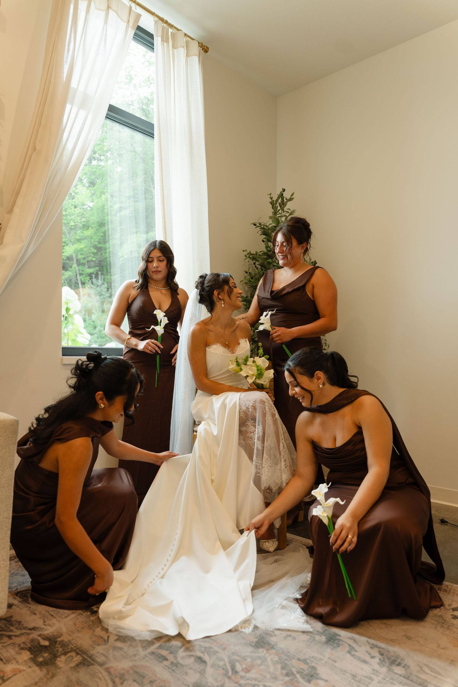 Bridesmaids in brown dresses arranging bride's white gown train near bright window.