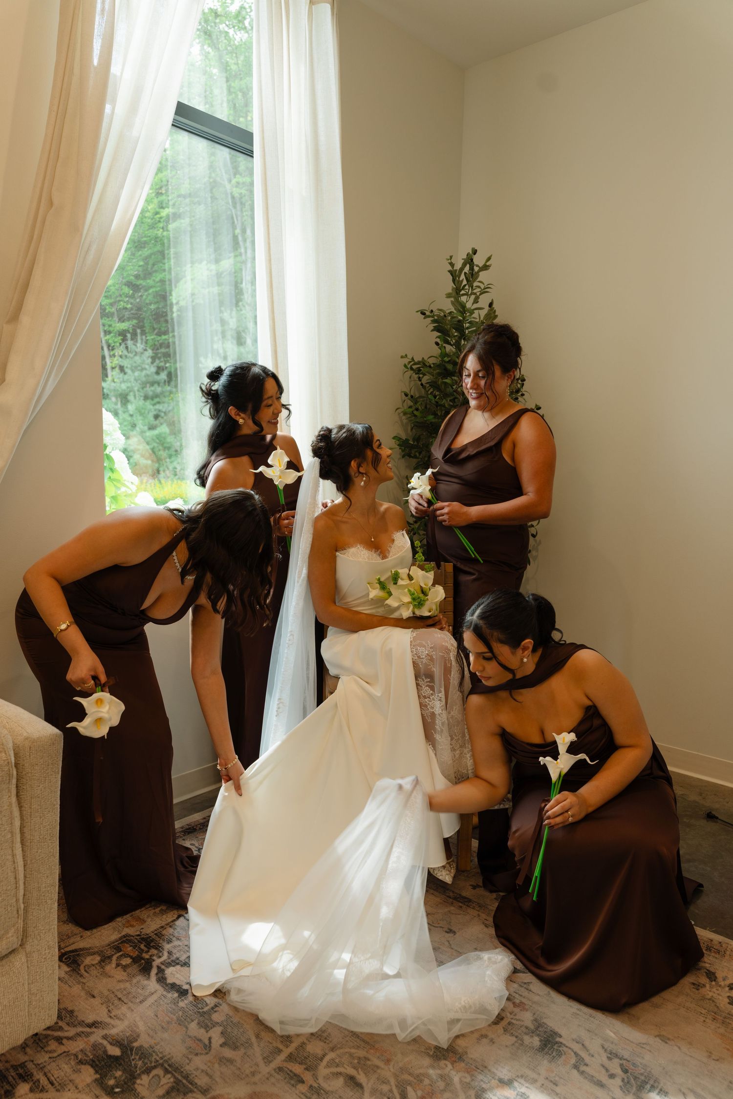 Bride in white gown surrounded by bridesmaids in brown dresses near draped window.