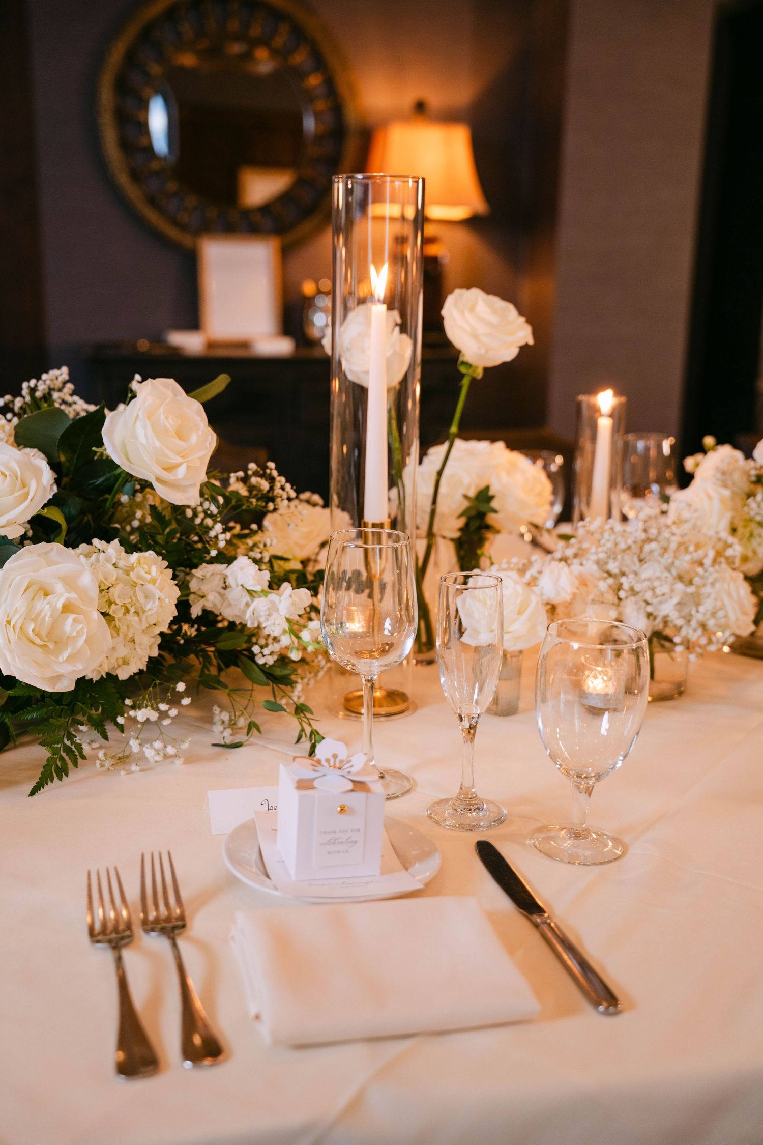 Elegant wedding reception table set with white roses, tall candle centerpiece, and gold accents.