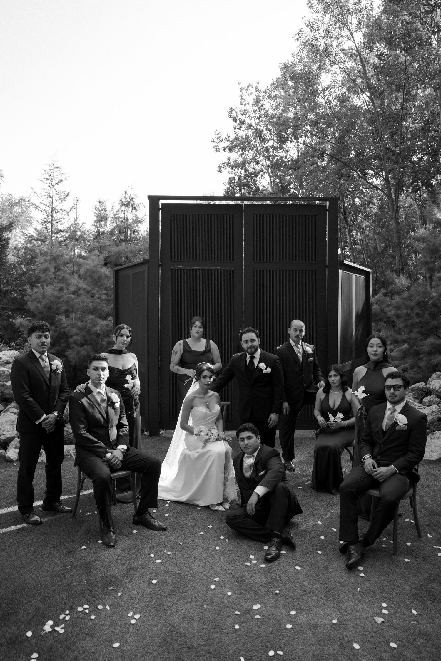 Black and white wedding party portrait posed in front of dark chapel structure surrounded by trees.