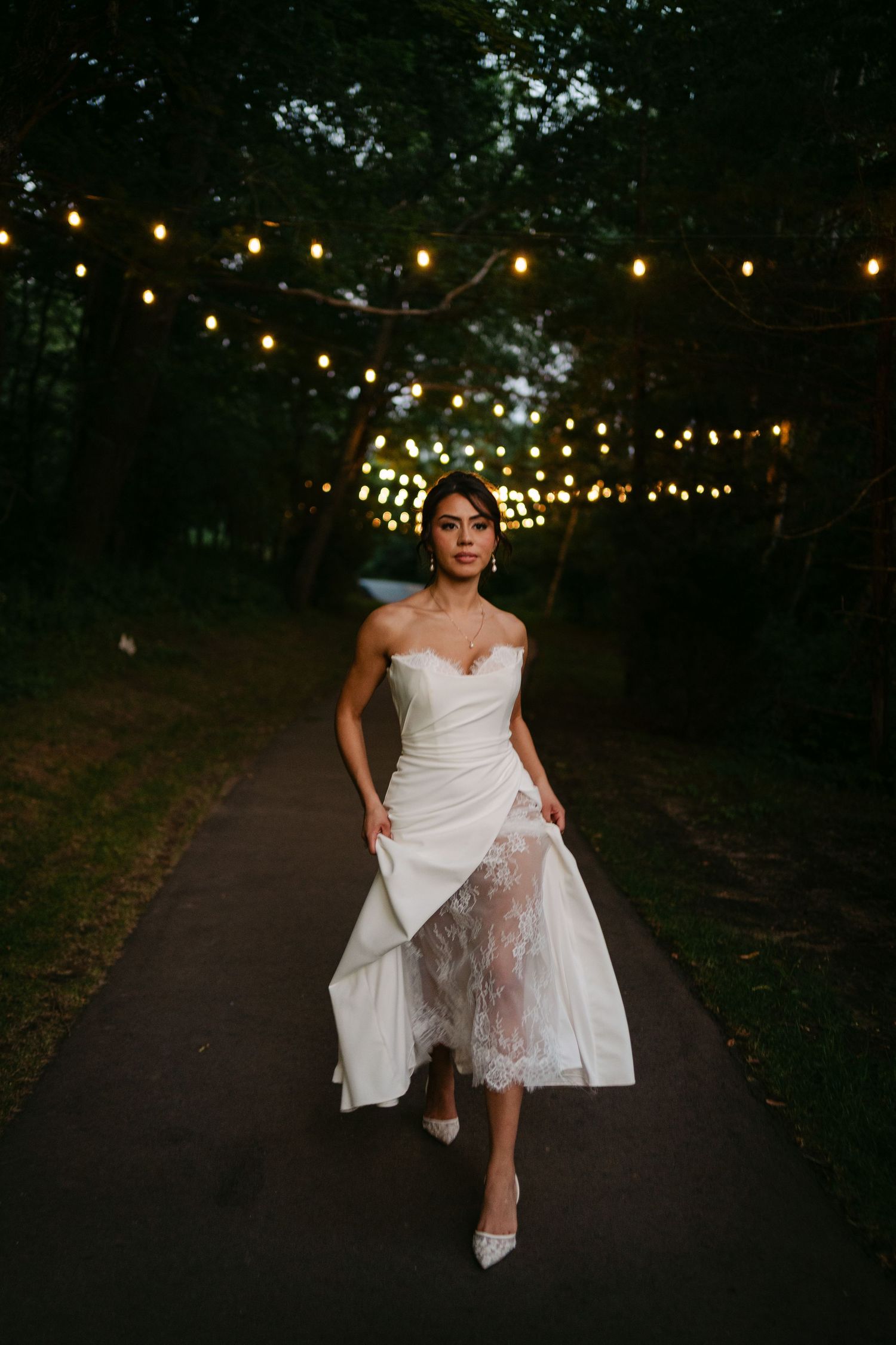 Beautiful bride in strapless white wedding gown with lace overlay walks along illuminated garden path.