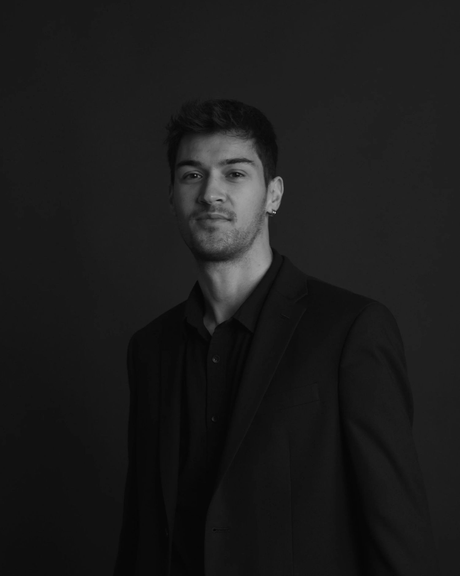 Black and white portrait of a young man in a dark blazer posing against a dark studio background.