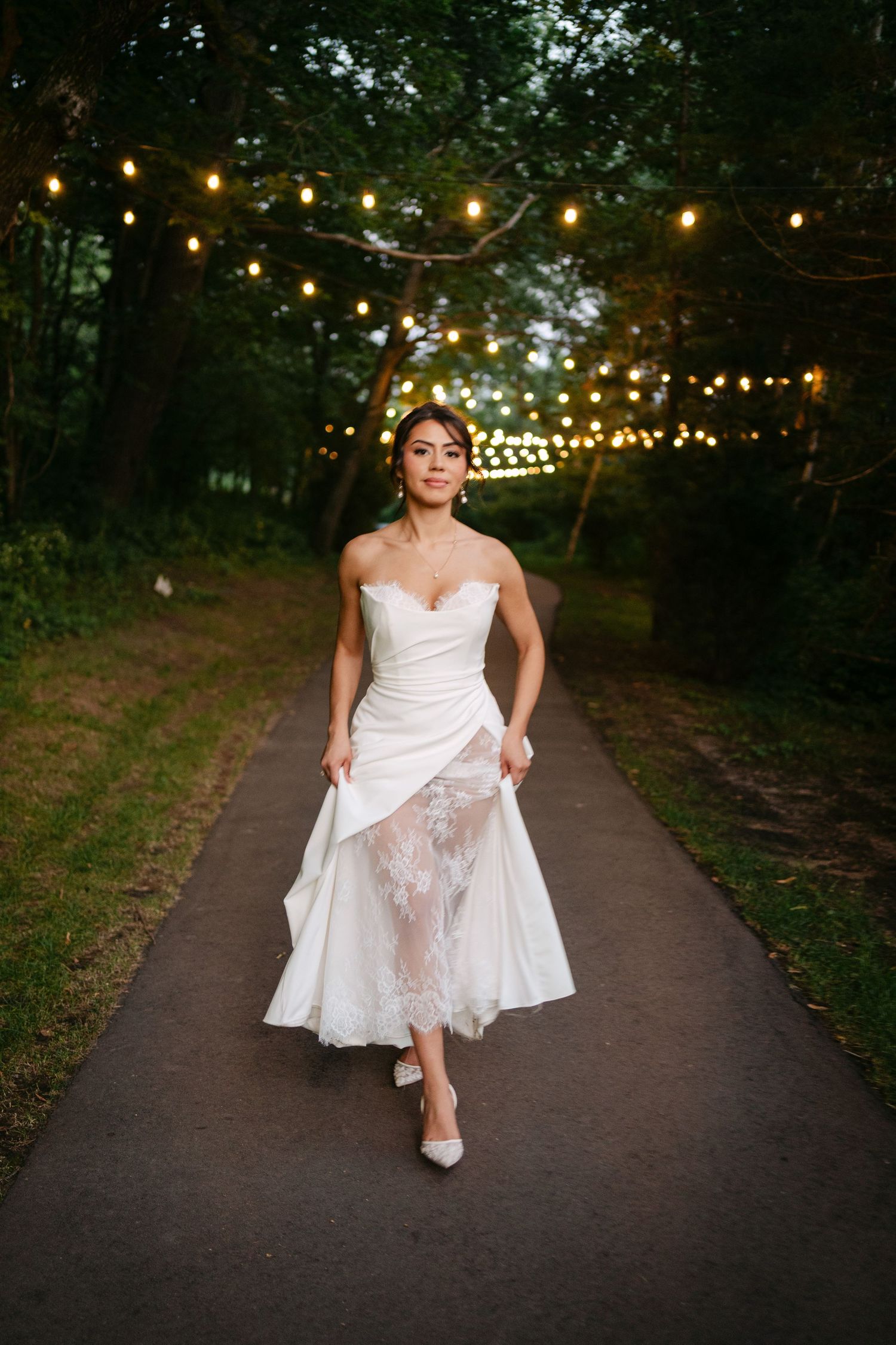 Bride in white strapless lace wedding dress walks down tree-lined path with string lights at dusk.