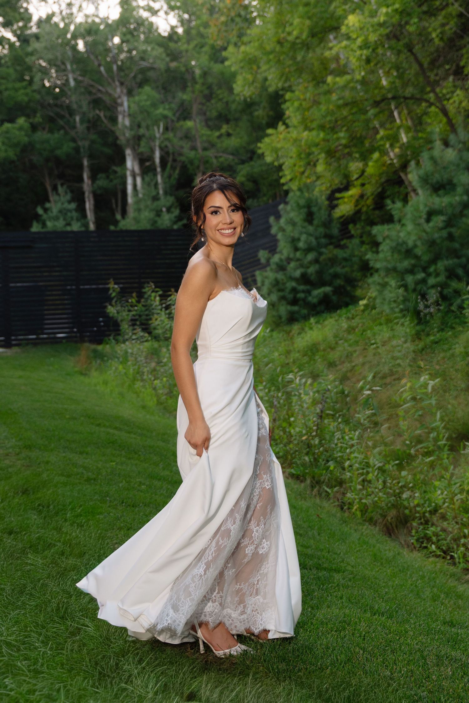 Radiant bride twirls in a strapless white lace wedding gown on a lush green lawn at golden hour.