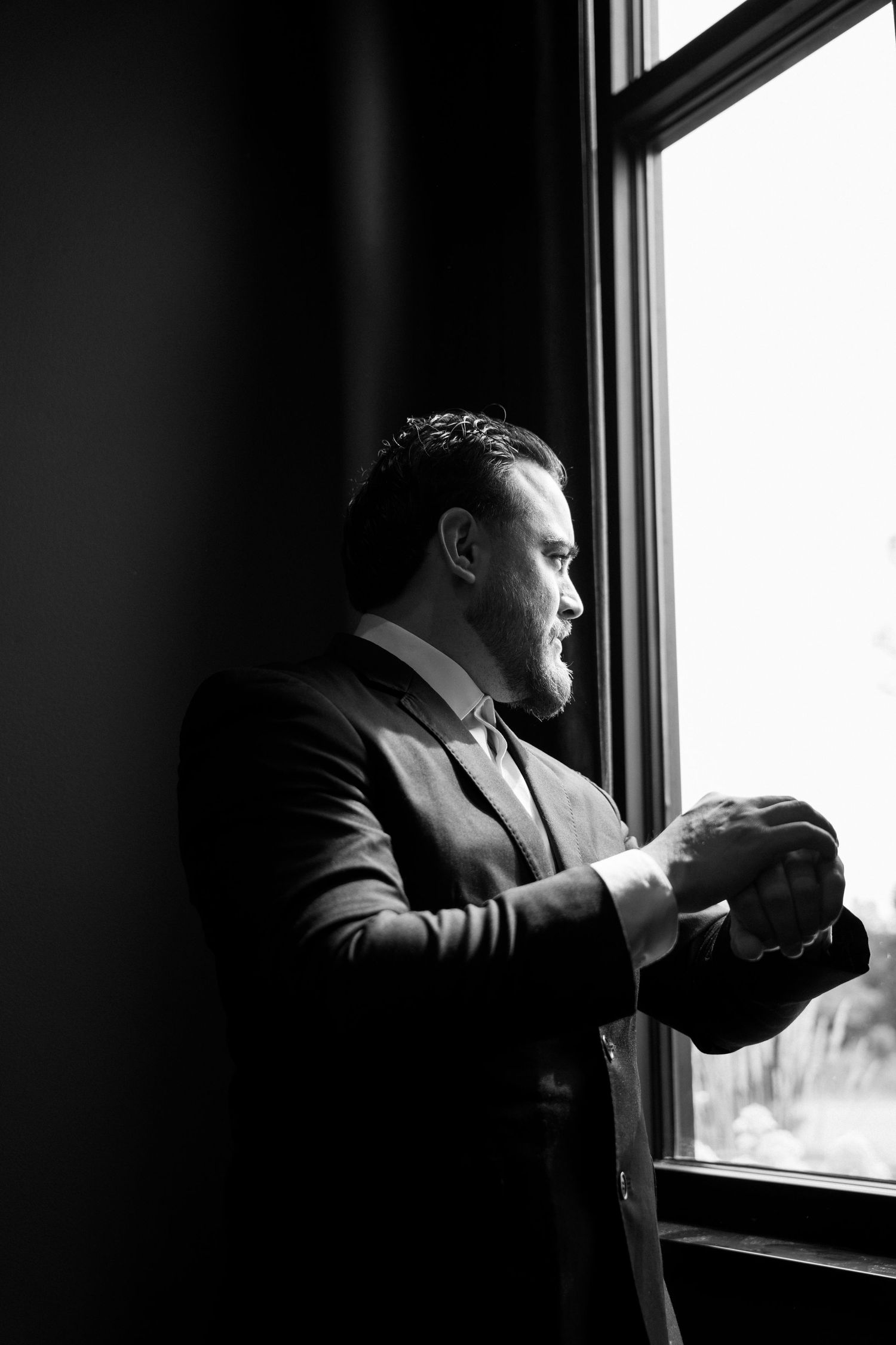 Black and white photo of groom in dark suit adjusting cufflinks while standing by a bright window.