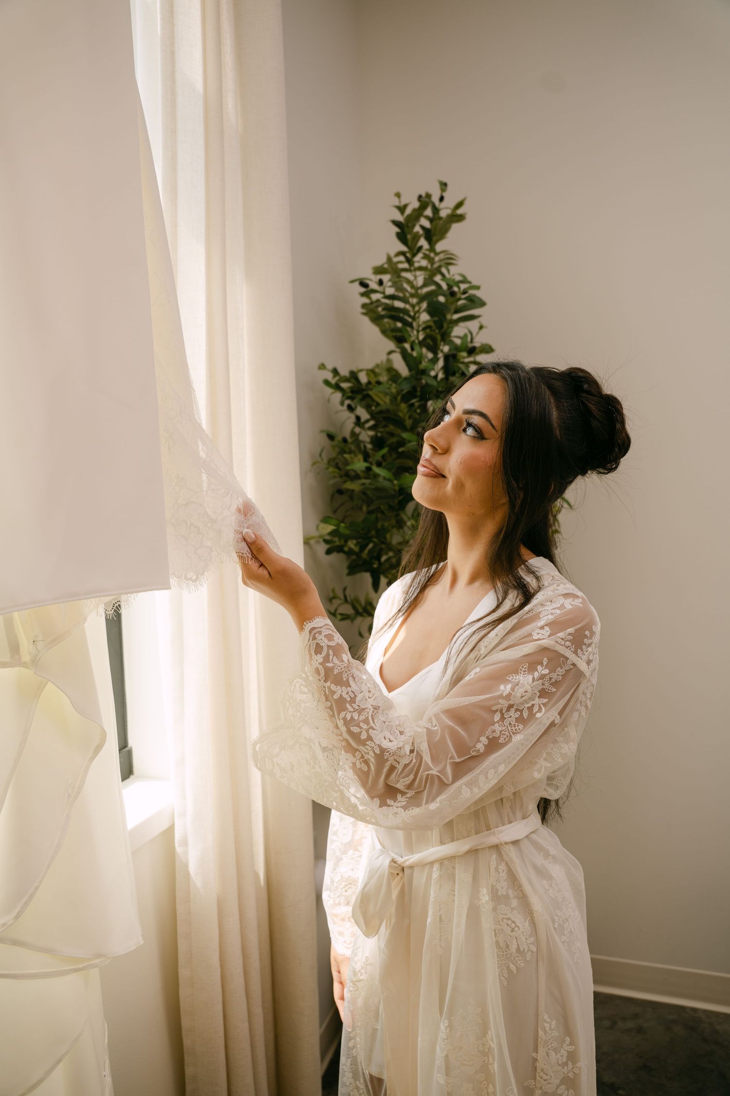 Bride in white lace robe gazing at wedding dress hanging by sunlit window with green plant backdrop.