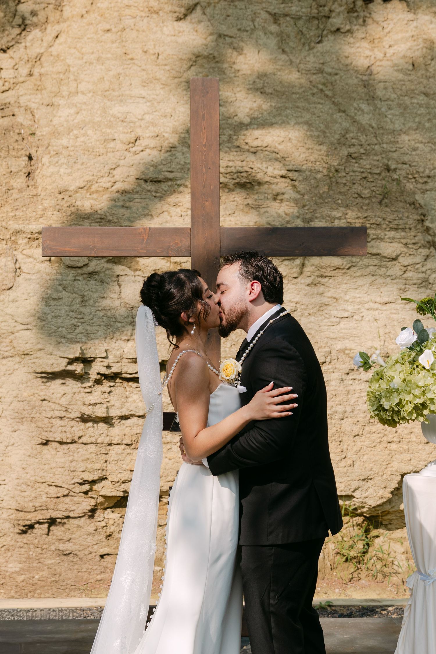 Bride and groom share first kiss at altar with wooden cross against stone wall backdrop.