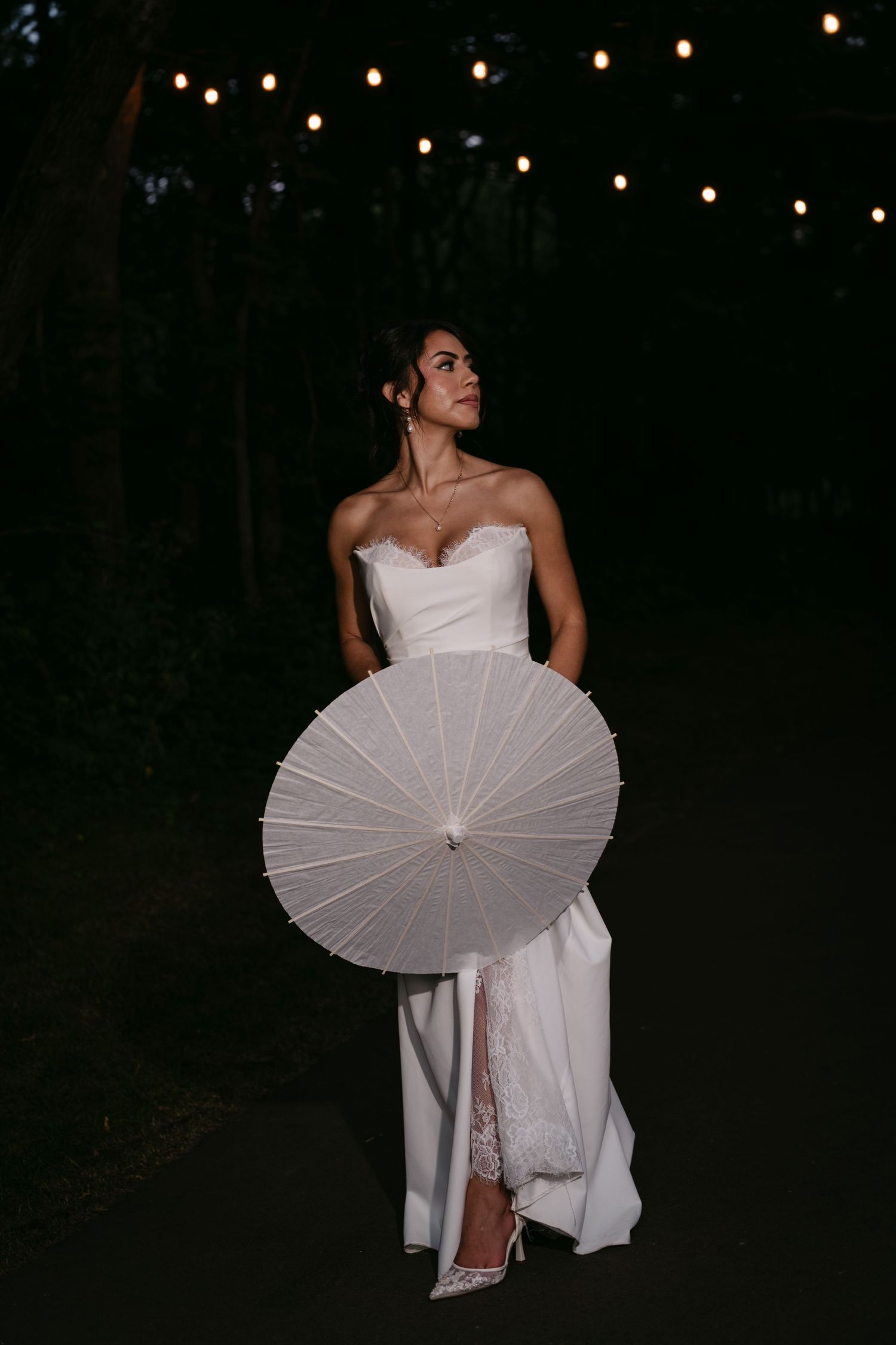 Bride in strapless white lace gown holding parasol gazes upward on dark tree-lined pathway.