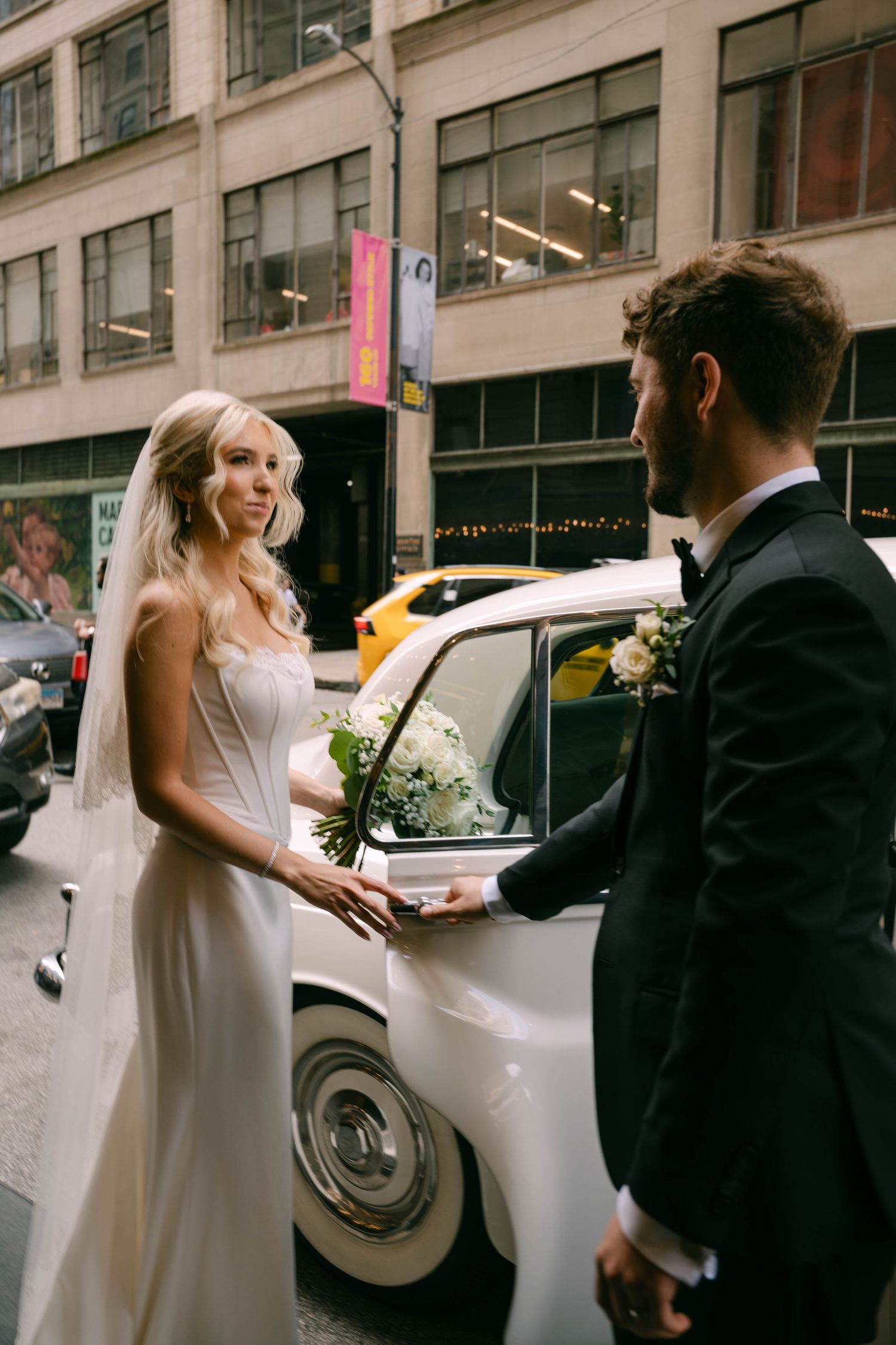 Bride and groom standing by vintage white car on busy city street, bride holding white bouquet.