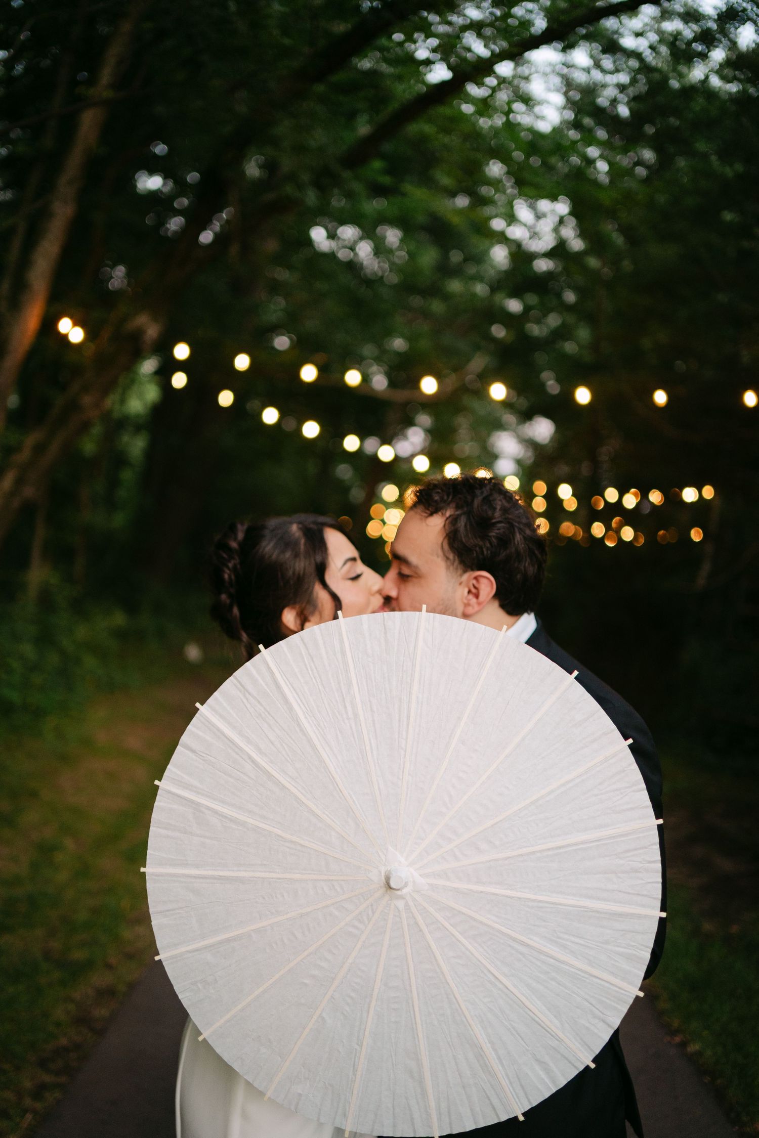 Bride and groom share a kiss hidden behind a white paper parasol on a garden path.